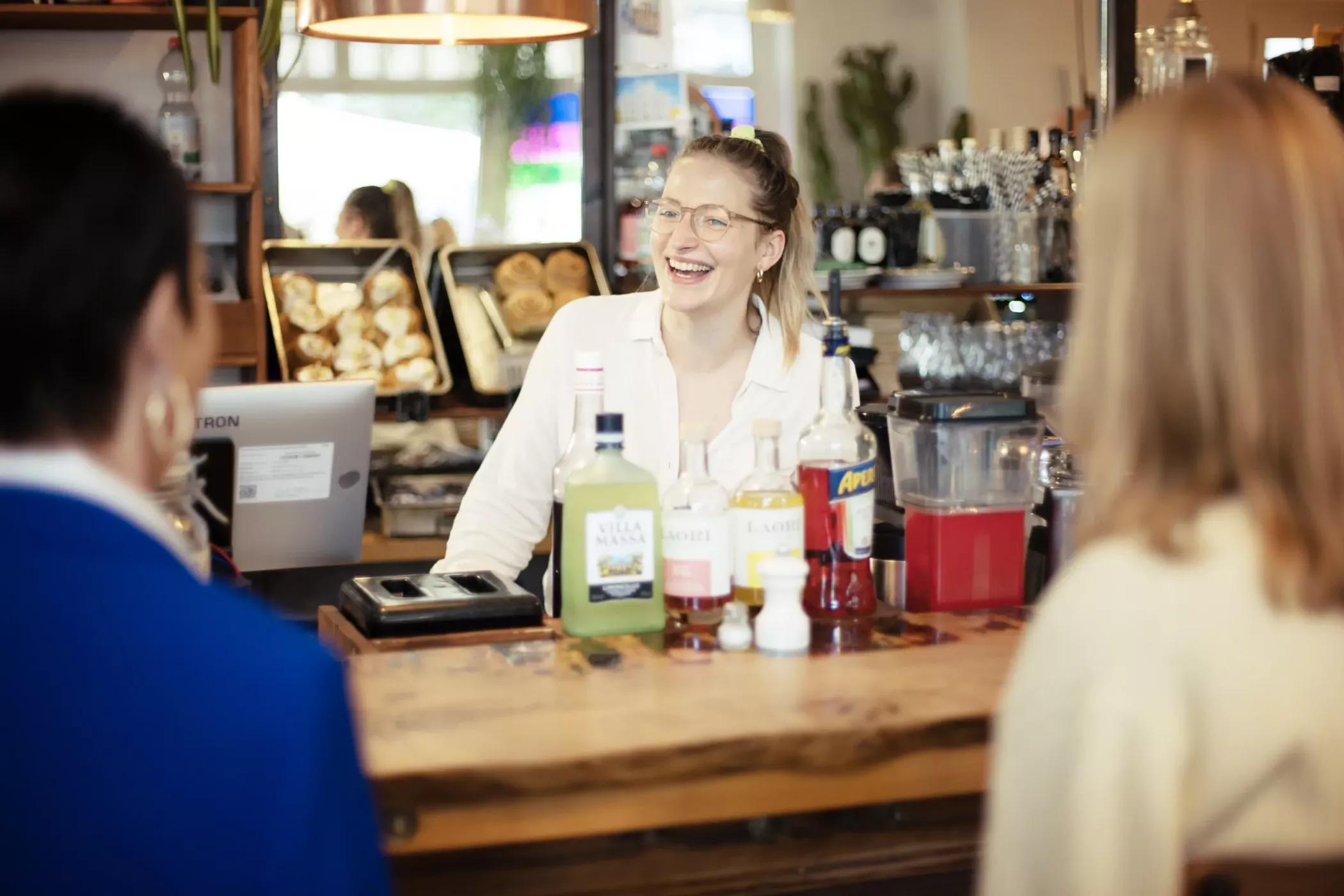 A smiling waiter stands behind a wooden counter and talks to two customers in a cozy café.