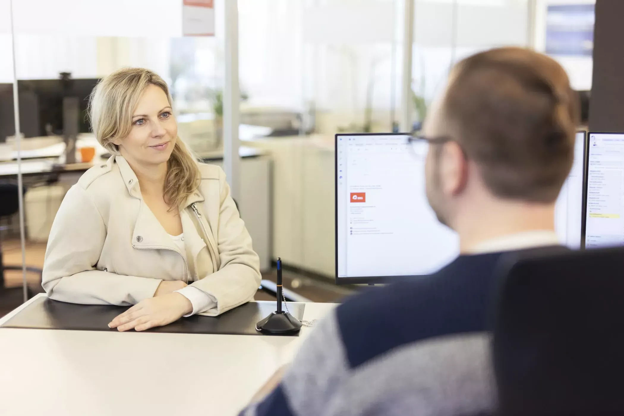 Eine Frau, die an einem Schreibtisch sitzt, gegenüber einem Mann mit einem Computerbildschirm, der ein Gespräch in einem modernen Büro führt.