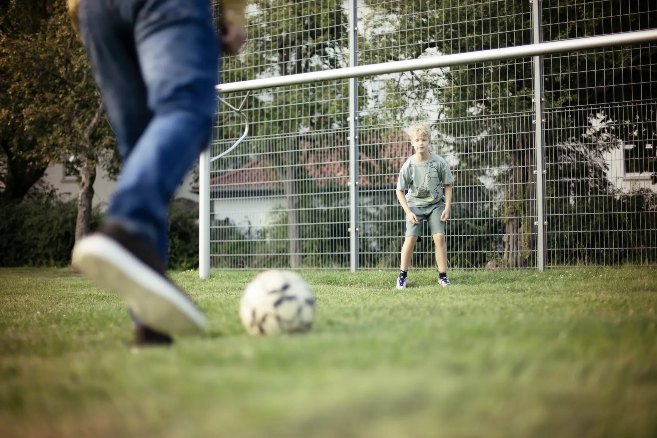 Ein Kind steht vor einem Tor auf einer Wiese und bereitet sich darauf vor, einen von einem Erwachsenen getretenen Fußball zu stoppen.