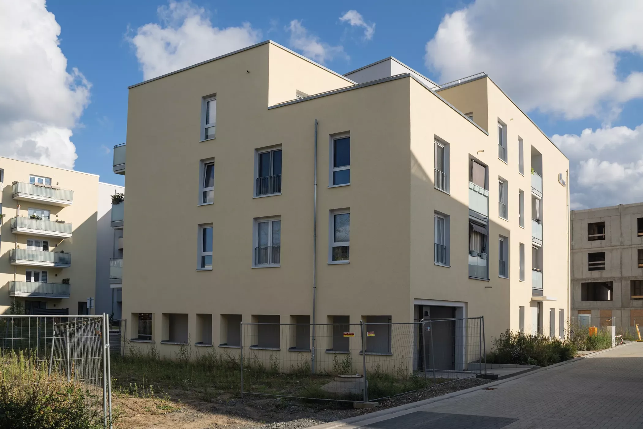 A modern beige-colored residential building with several windows, balconies and a fenced-in area under a partly cloudy sky.