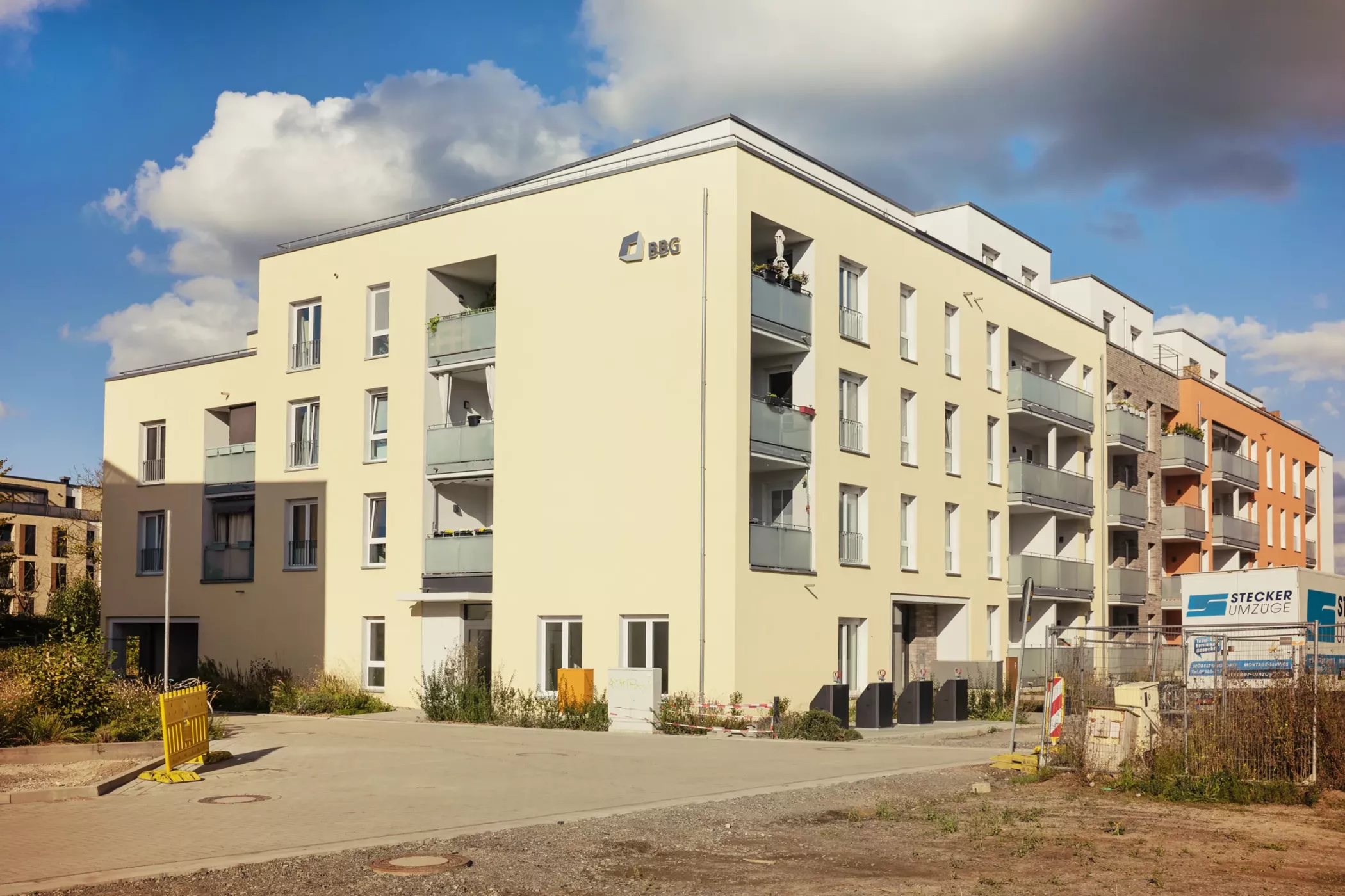 Ein modernes beigefarbenes Mehrfamilienhaus mit Balkonen, unter teilweise bewölktem Himmel, neben einer Baustelle mit Absperrungen und Schildern.