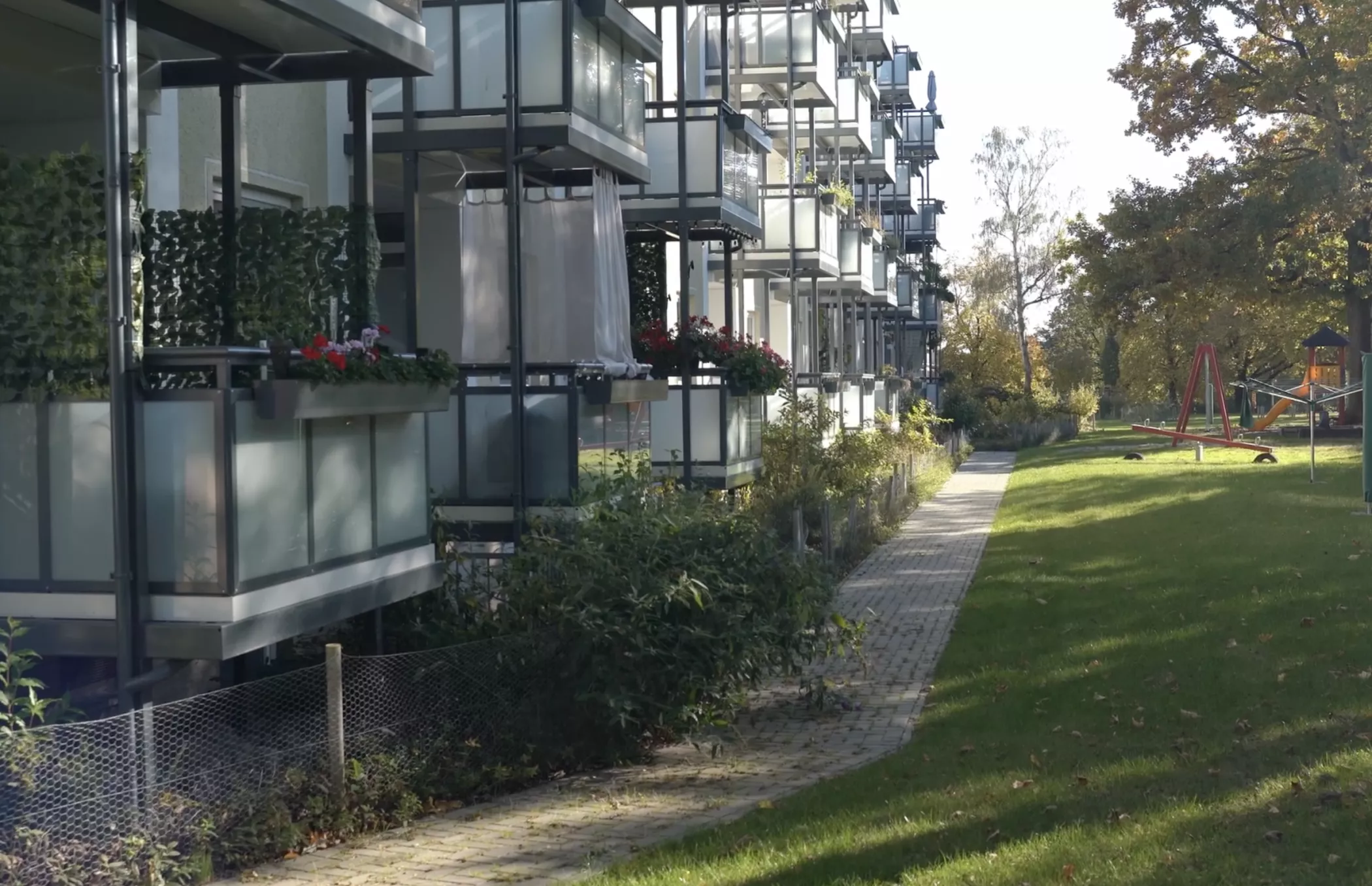 A modern residential building with glass balconies, plants and a walkway next to a green area on a sunny day.