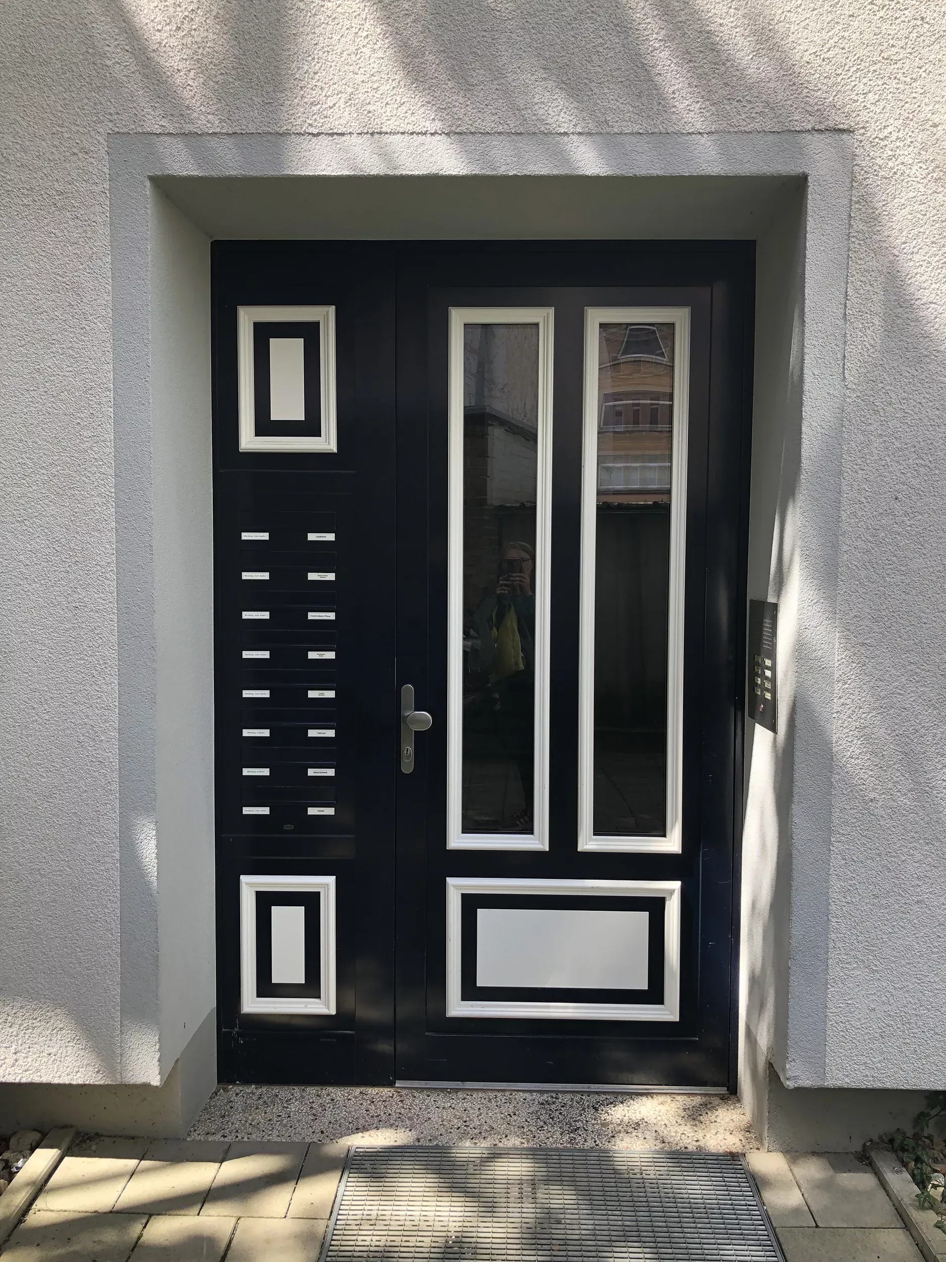 Porte moderne noire avec des motifs géométriques blancs et des vitres blanches sur un mur gris clair avec quelques ombres visibles.
