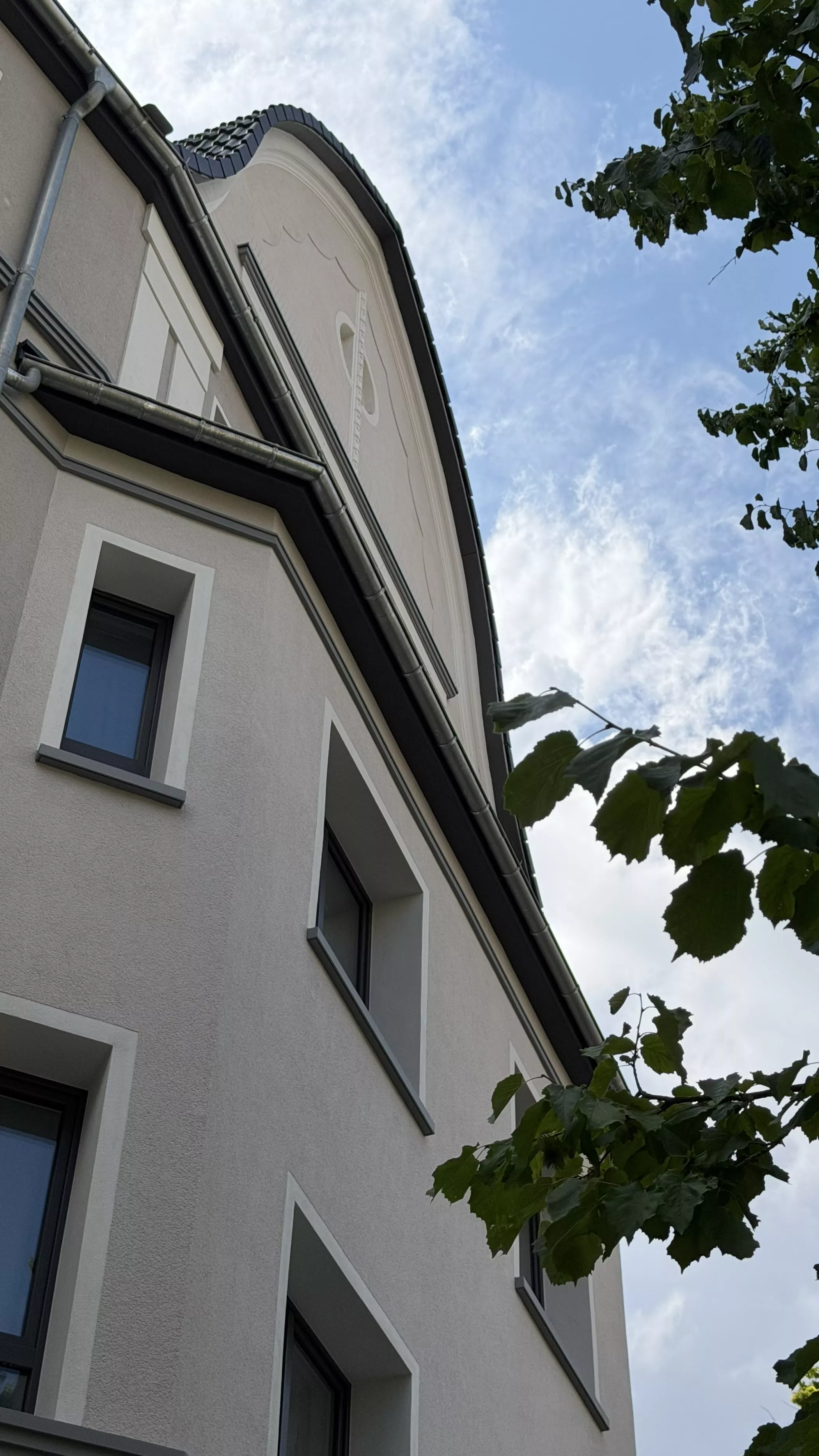 Ein beiges Gebäude mit weißen Rändern, von unten gesehen, neben üppig grünen Zweigen unter einem teilweise bewölkten Himmel.