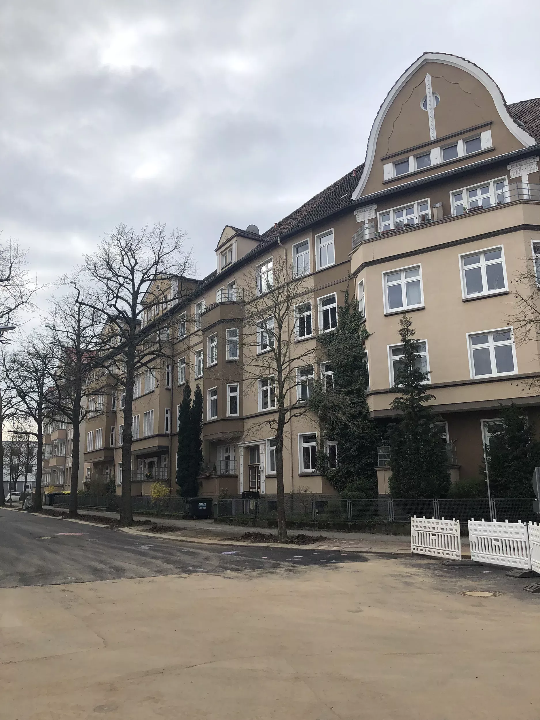Ein mehrstöckiges, beigefarbenes Wohnhaus mit weißen Zierleisten steht an einer ruhigen, leeren, von Bäumen gesäumten Straße unter einem bedeckten Himmel.