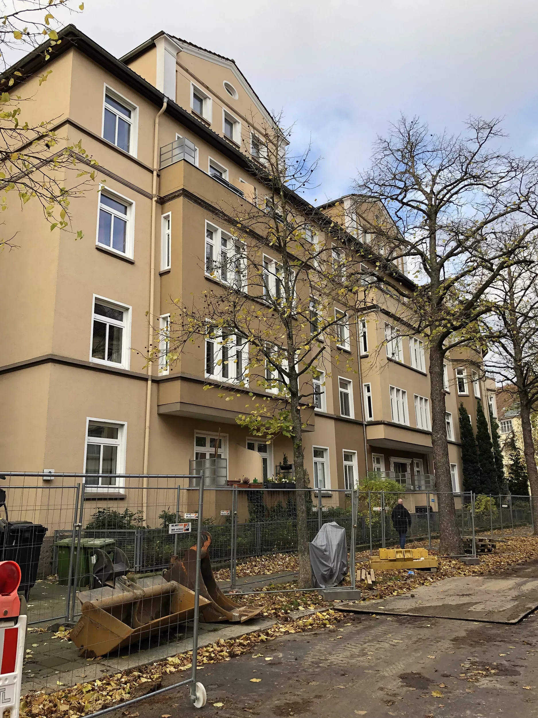 Ein vierstöckiges beigefarbenes Wohnhaus mit kahlen Bäumen und Baugeräten hinter einem Metallzaun an einem bewölkten Tag.