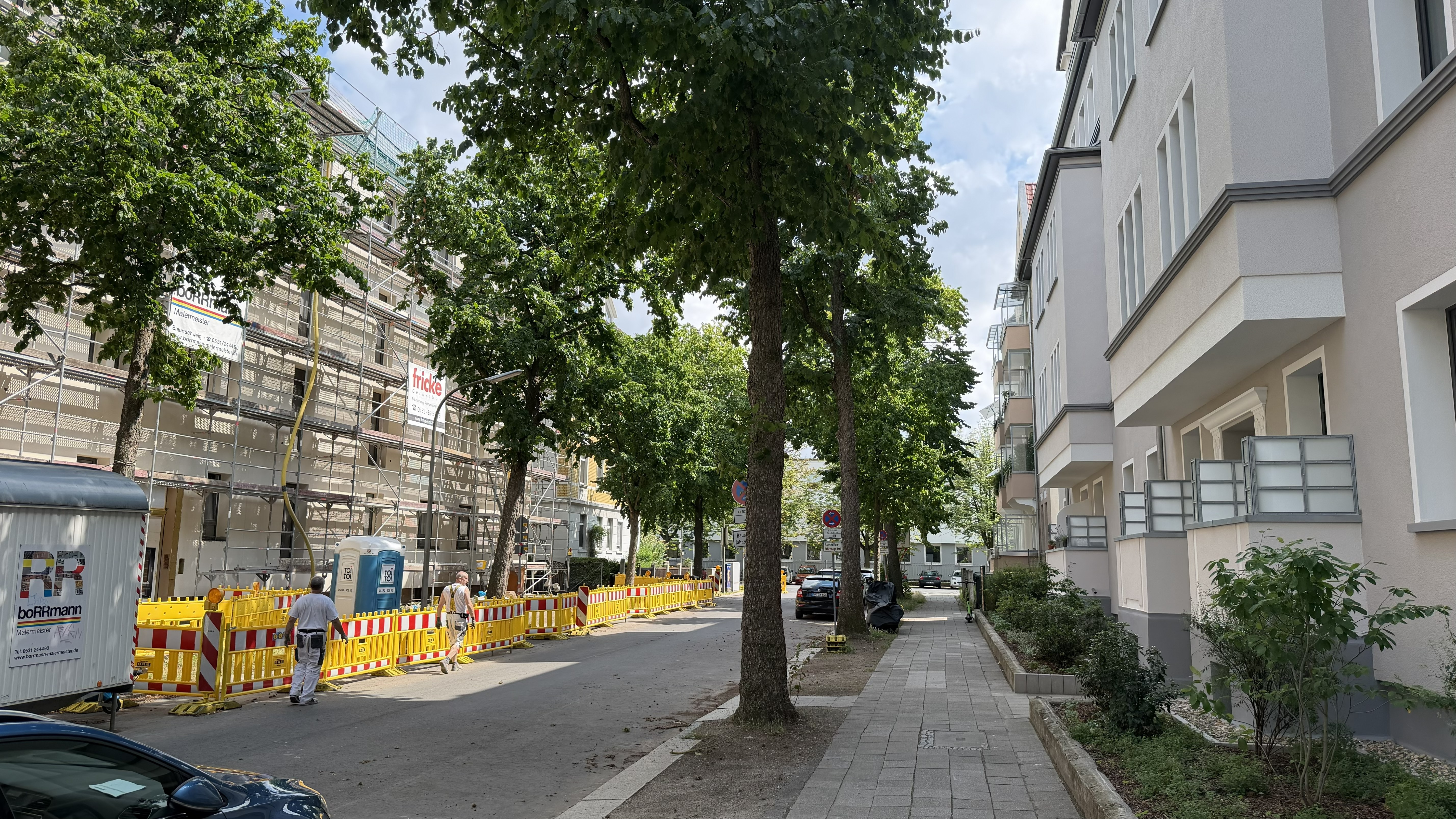 Eine Straße in der Stadt, auf deren linker Seite Bauzäune stehen, an deren beiden Seiten Bäume stehen und auf deren beiden Seiten Wohnhäuser stehen.