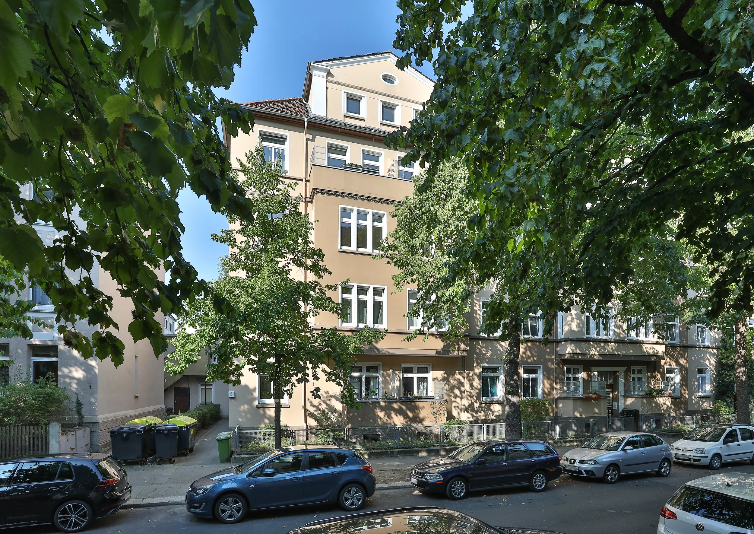 Ein beigefarbenes Wohnhaus mit weiß gestrichenen Fenstern und Autos, die entlang der von Bäumen gesäumten Straße davor parken.