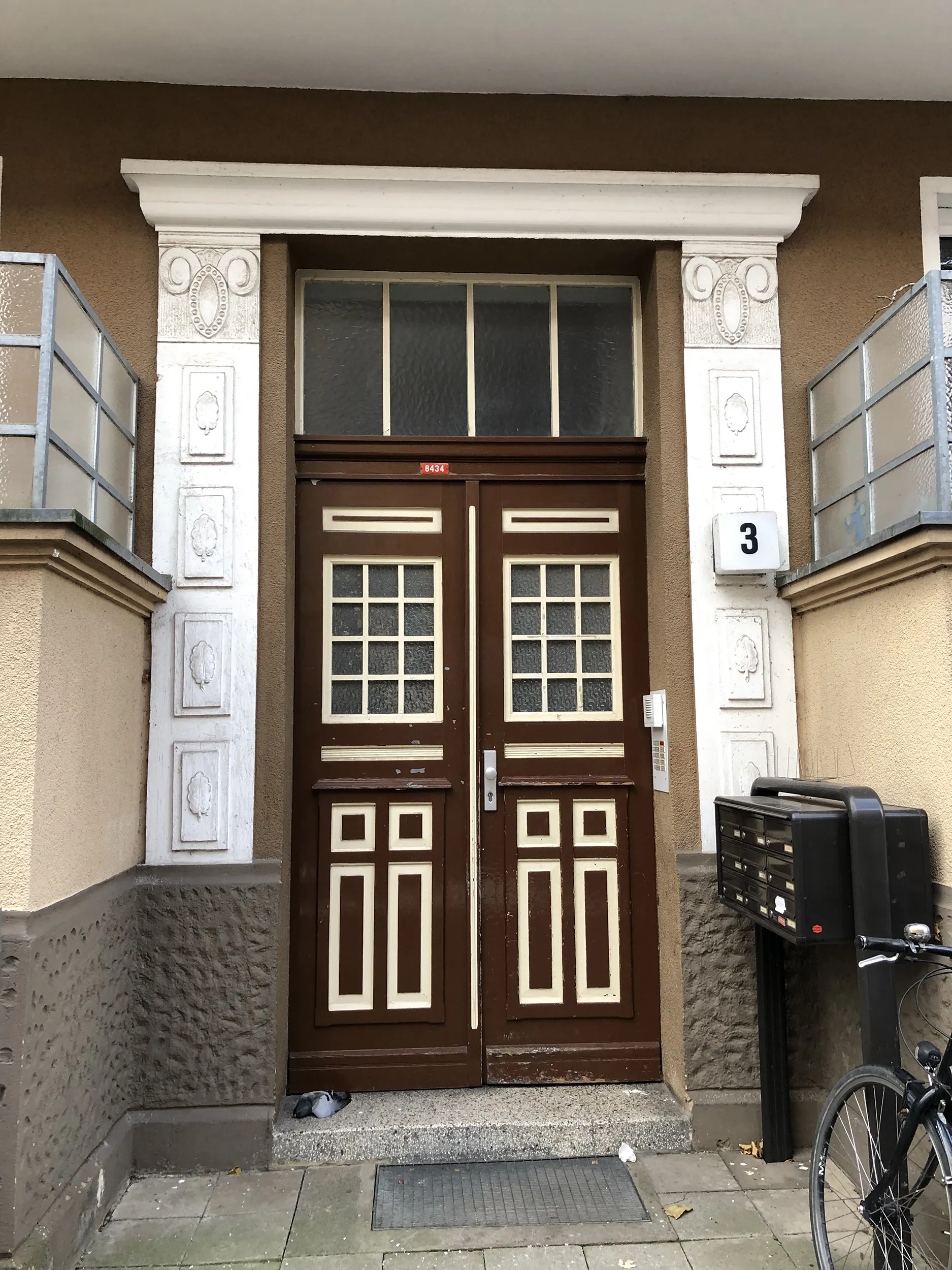 Doppelte Holztüren mit Glasscheiben, flankiert von Briefkästen und einem Fahrrad am Eingang zu einem braunen Gebäude mit weißer Verkleidung.