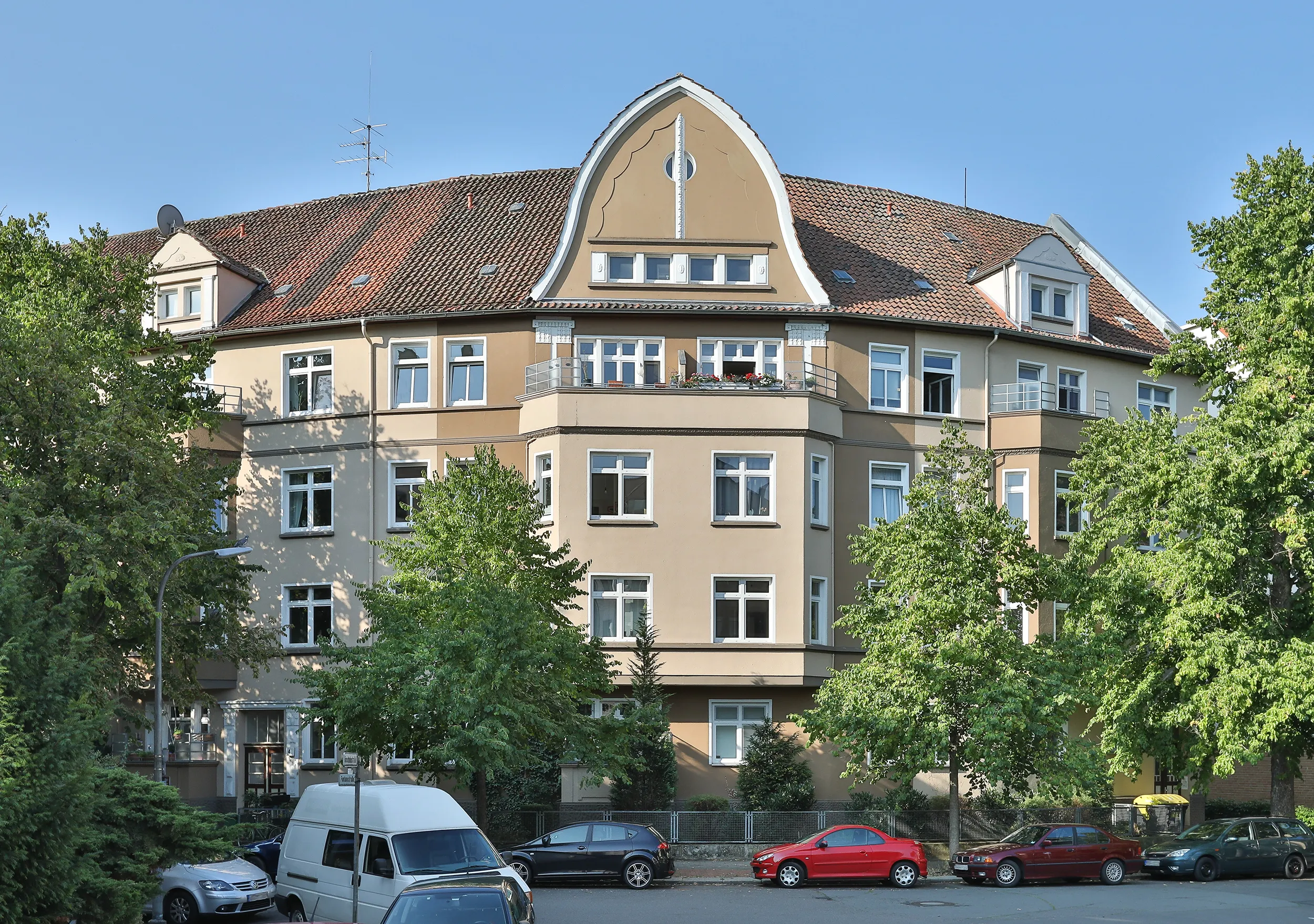 Ein braunes Mehrfamilienhaus mit rotem Ziegeldach, einem geschwungenen Giebel in der Mitte und parkenden Autos entlang der von Bäumen gesäumten Straße davor.