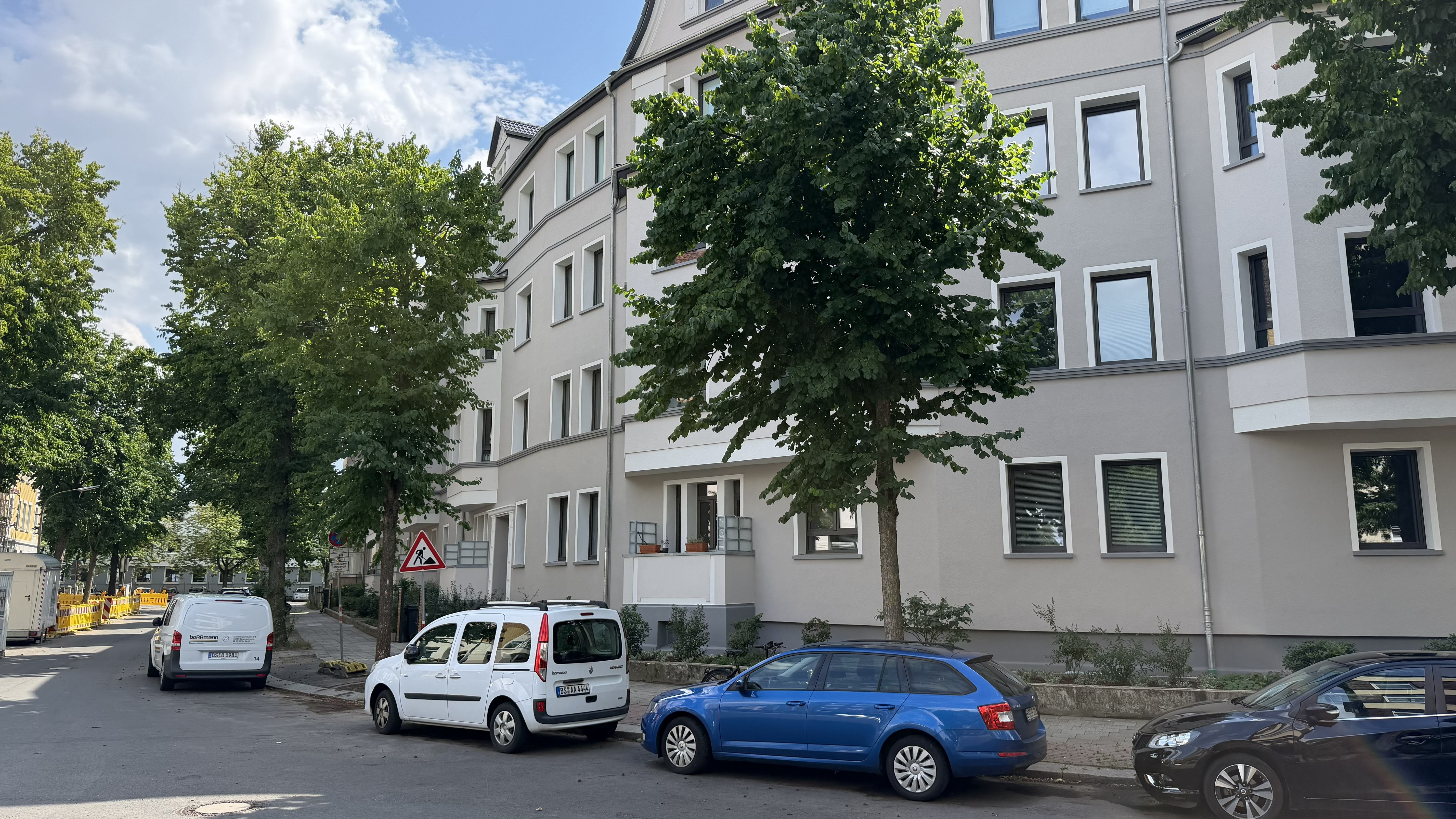 Drei Autos stehen in einer Straße vor einem hellgrauen Wohnhaus, und auf beiden Seiten des Bürgersteigs reihen sich Bäume aneinander.