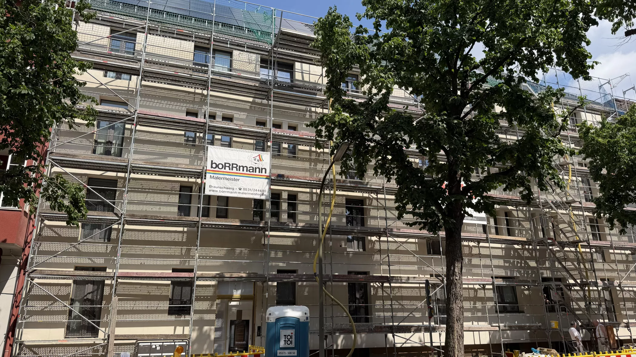 Ein Gebäude, das gerade renoviert wird, ist mit Gerüsten umgeben; davor stehen ein Baum und eine mobile Toilette.