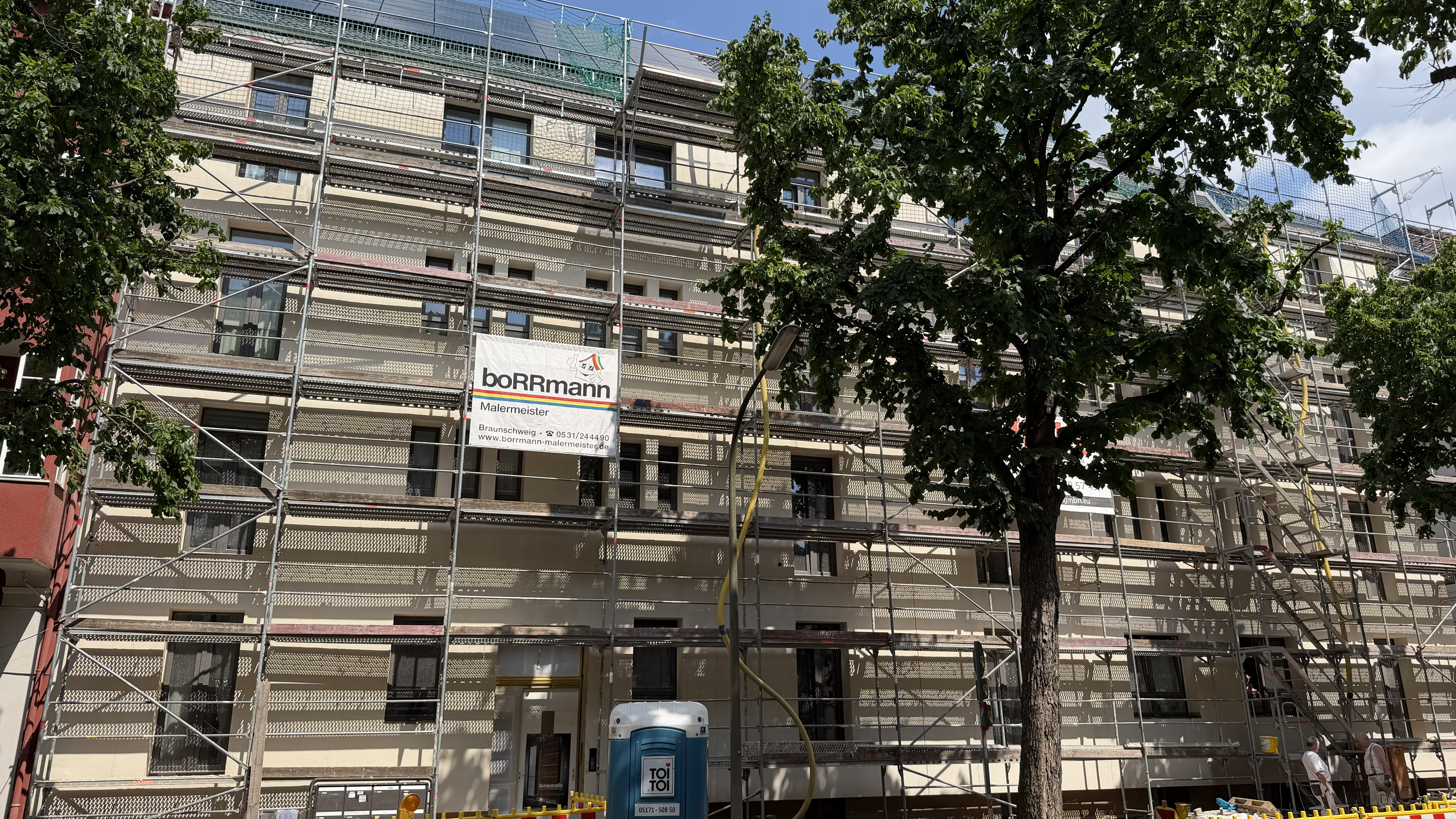Ein Gebäude, das gerade renoviert wird, ist mit Gerüsten umgeben; davor stehen ein Baum und eine mobile Toilette.