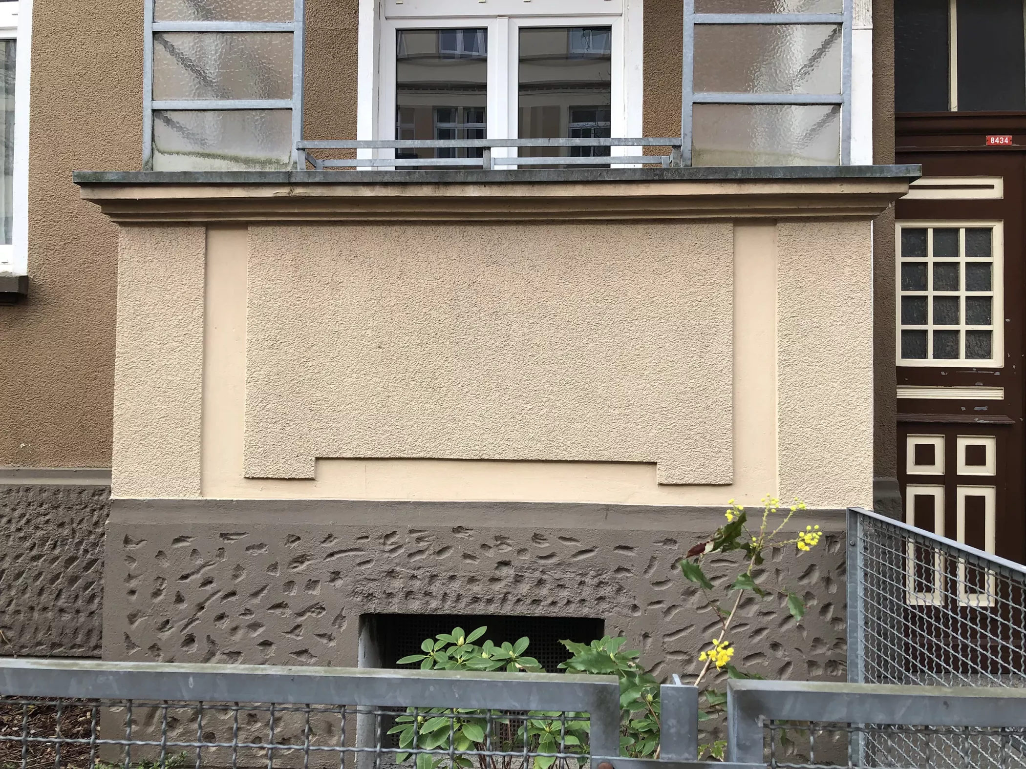 Eine beige gemusterte Wand mit einem kleinen Fenster am unteren Ende, gelbe Blumen und ein Metallzaun vor dem Eingang des Gebäudes.