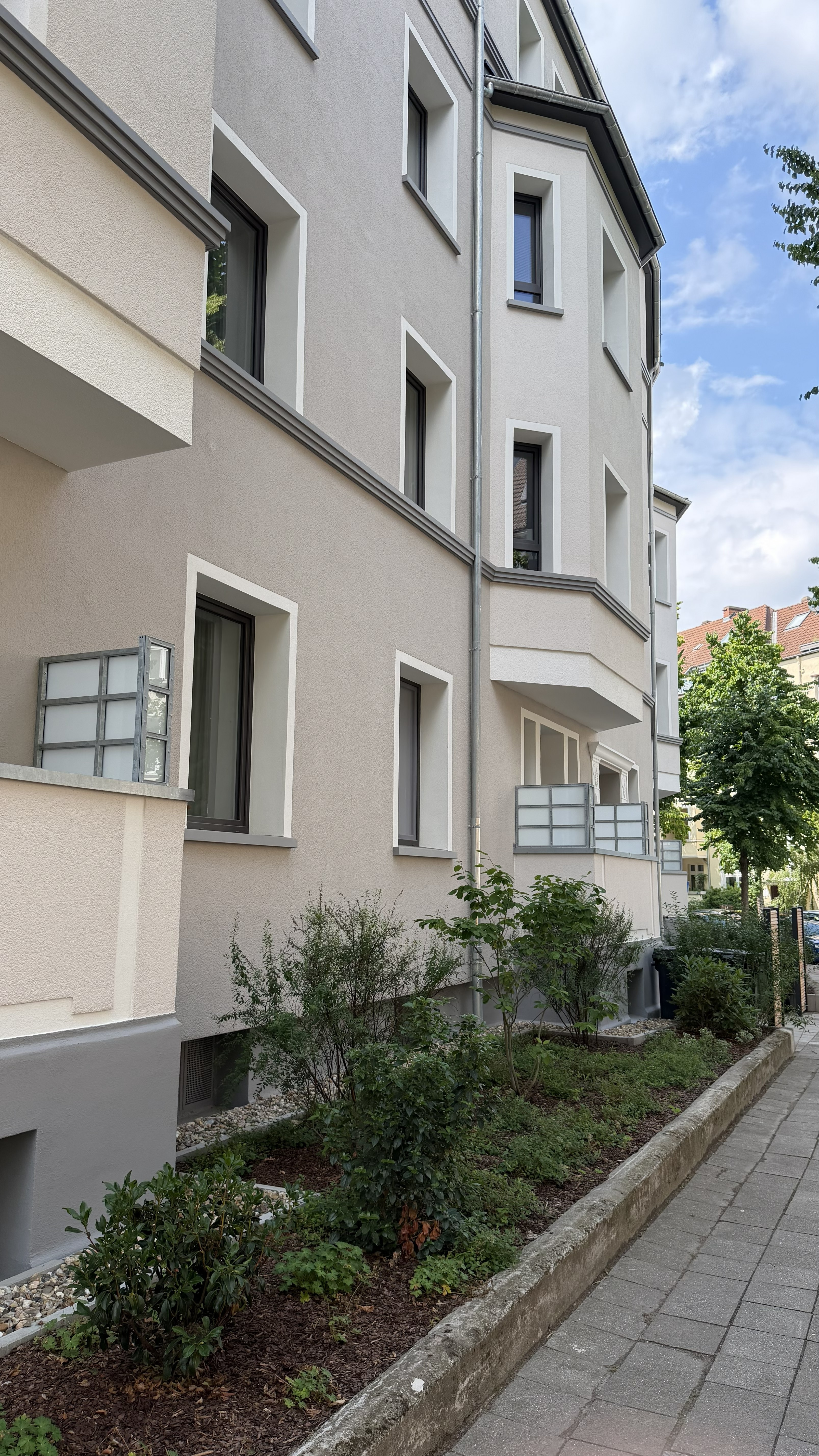 Ein modernes, beigefarbenes Wohnhaus mit Balkonen, Pflanzen und einem gepflasterten Bürgersteig unter einem teilweise bewölkten Himmel.
