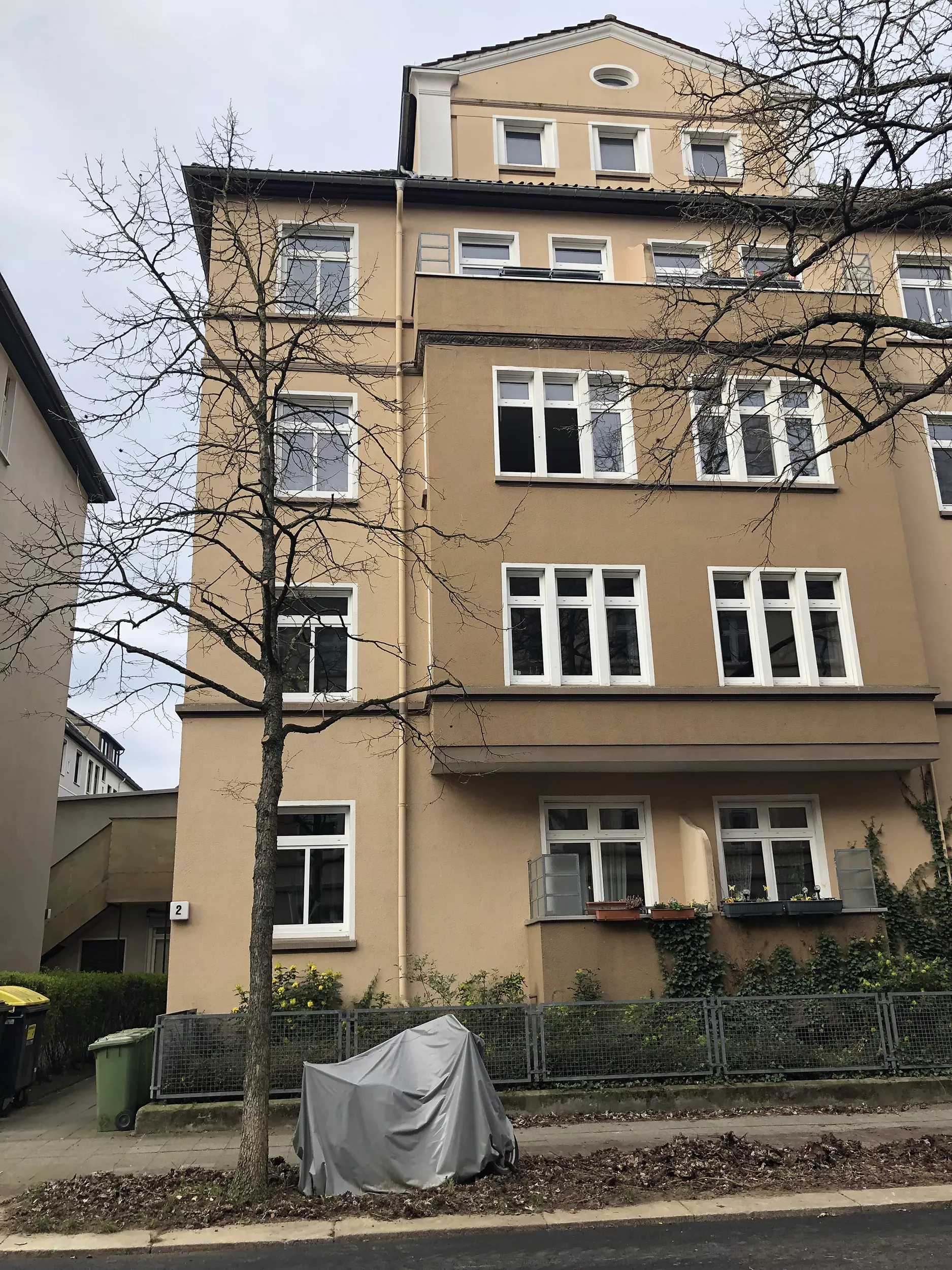 Ein beiges Wohnhaus mit weiß gerahmten Fenstern, blattlosen Bäumen und einem überdachten Motorrad, das auf der Straße geparkt ist.