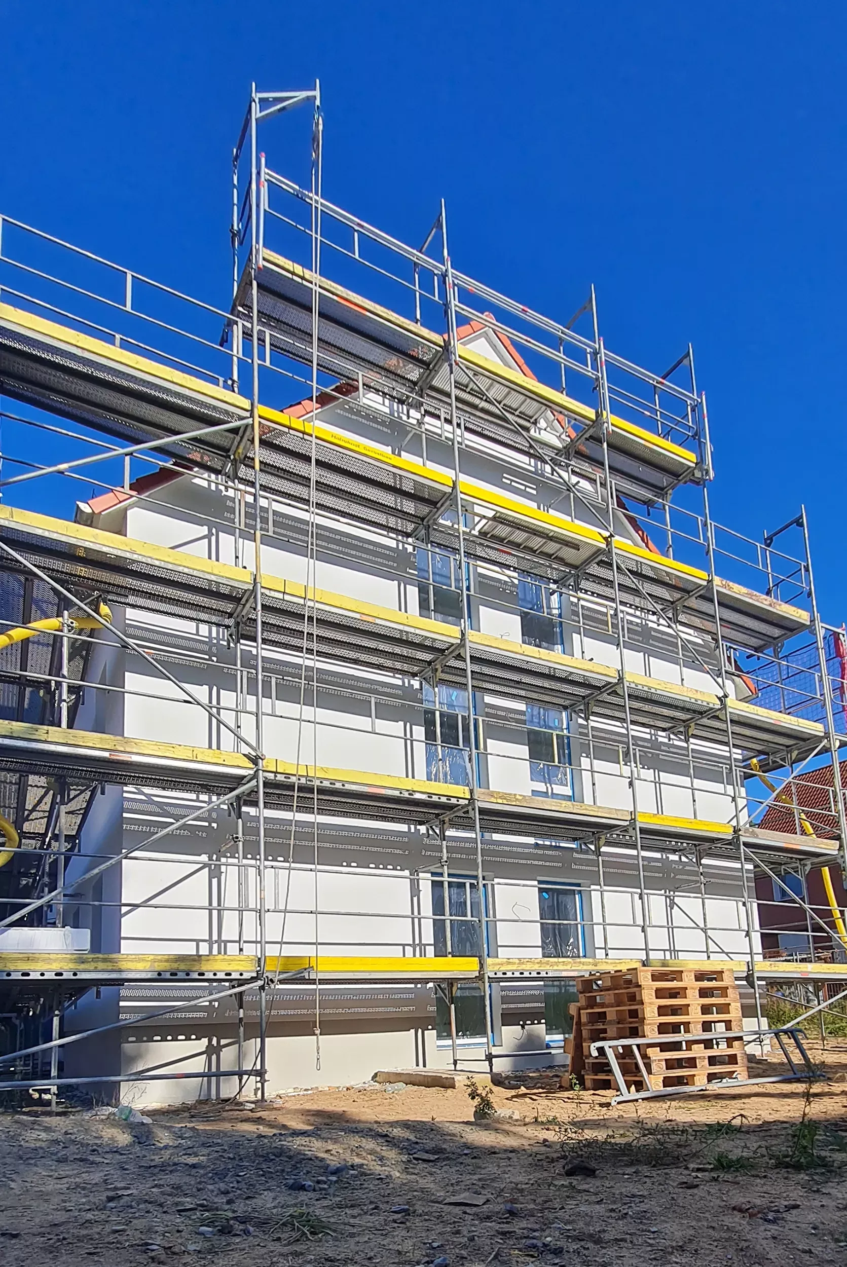 Ein weißes, im Bau befindliches Gebäude mit Gerüsten und Stapeln von Holzpaletten davor, unter einem klaren blauen Himmel.