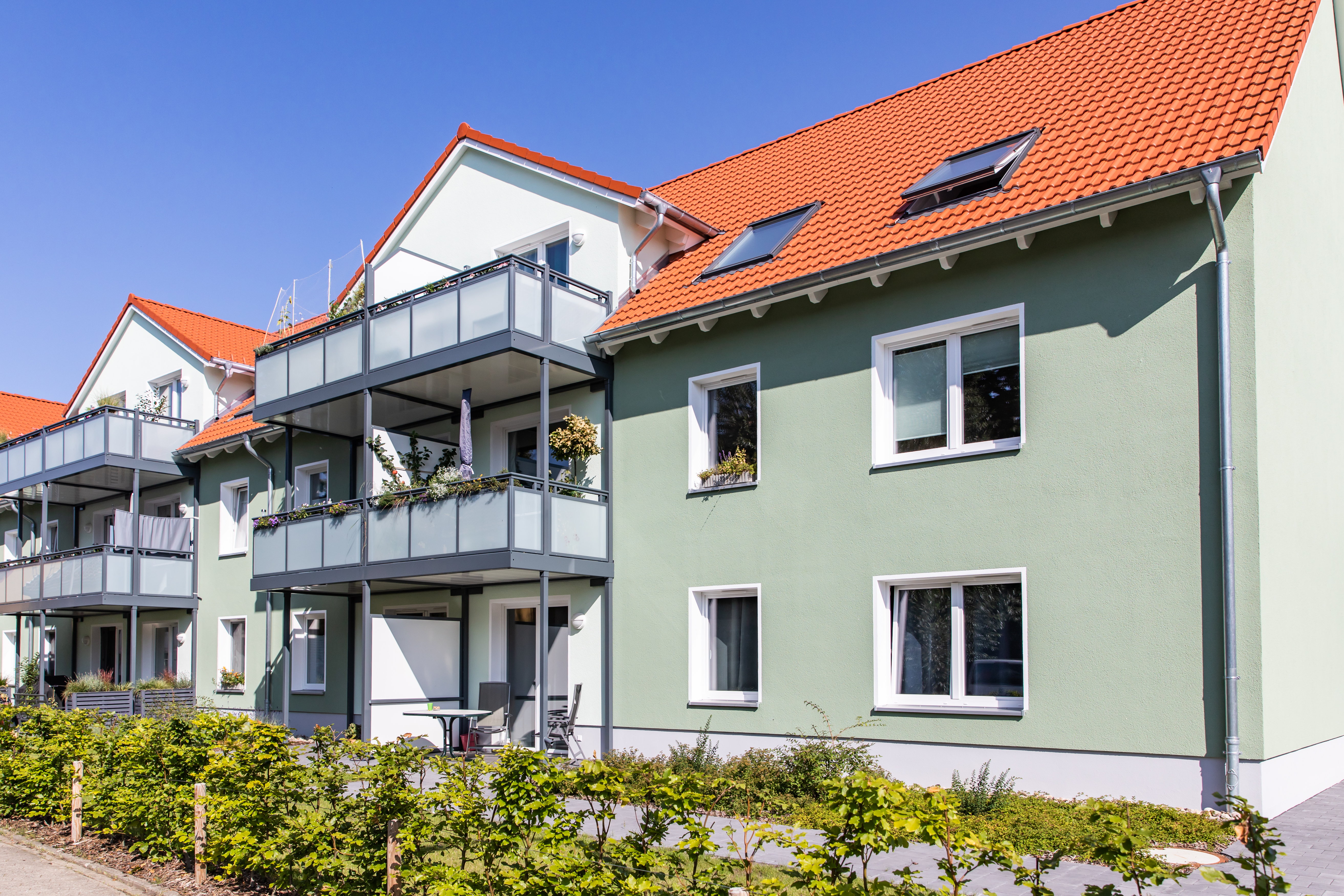 Ein modernes, hellgrünes, zeitgenössisches Wohngebäude mit rotem Dach, Balkonen und großen Fenstern vor einem klaren, blauen Himmel.