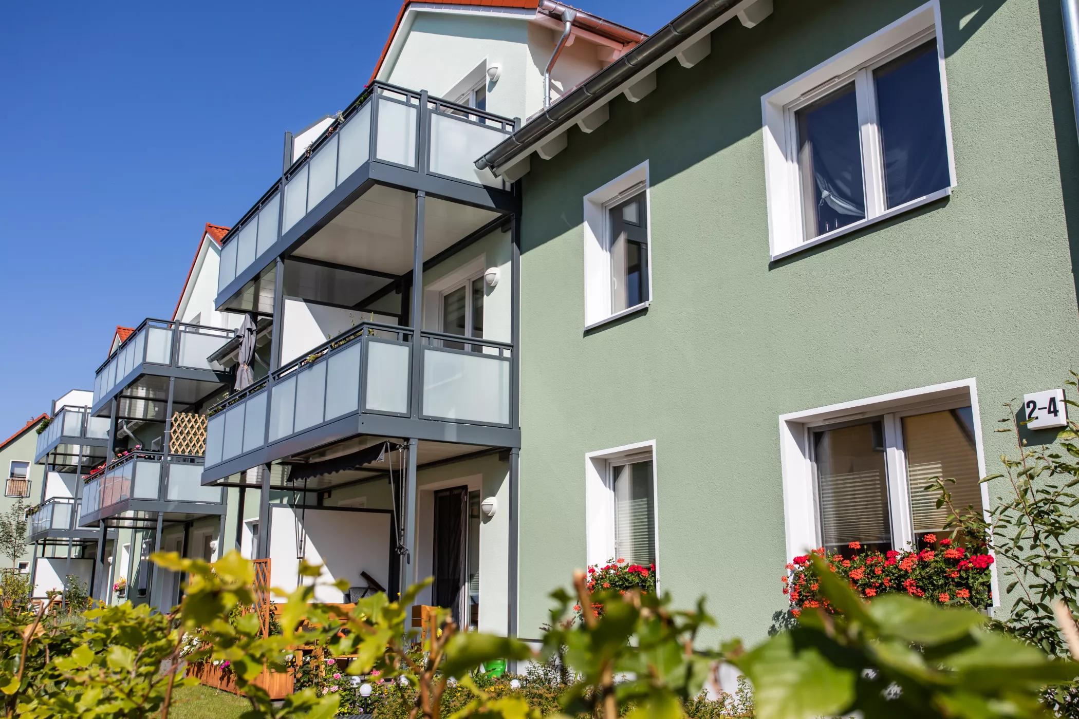 Ein modernes Wohngebäude, das sich durch begrünte Wände, verglaste Balkone und rote Blumen unter einem strahlend blauen Himmel auszeichnet.