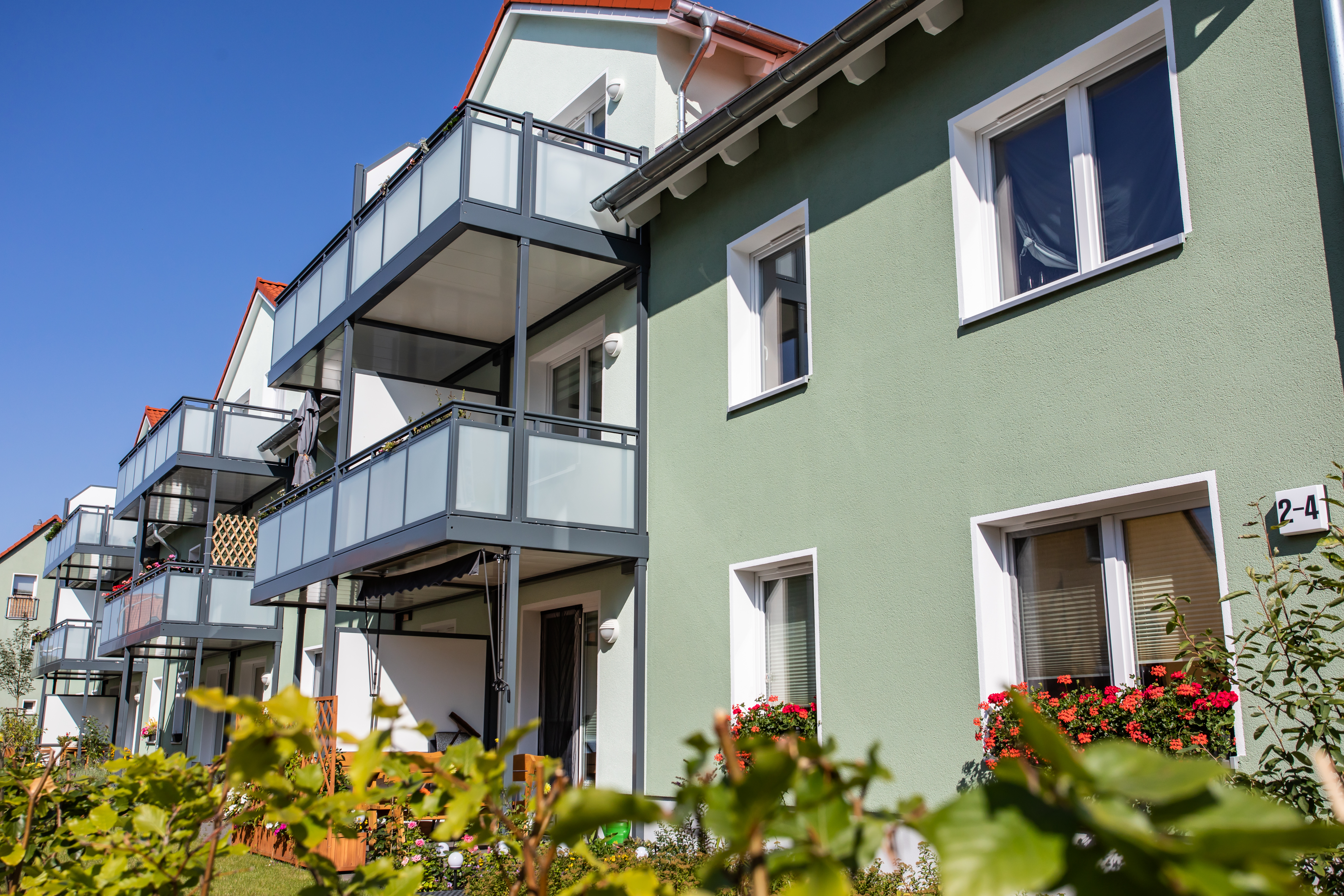 Ein modernes Wohngebäude, das sich durch begrünte Wände, verglaste Balkone und rote Blumen unter einem strahlend blauen Himmel auszeichnet.