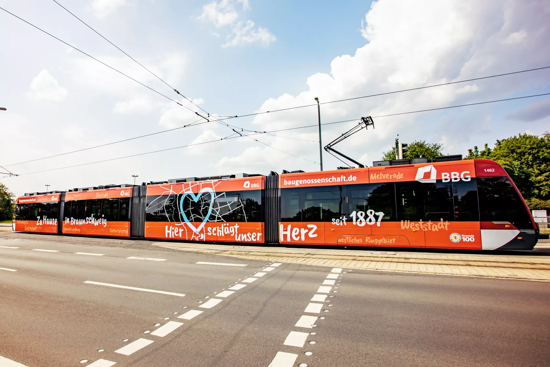 Eine leuchtend rote Straßenbahn mit weißem Schriftzug und Text parkt an einem sonnigen Tag an einer Kreuzung, mit Bäumen und blauem Himmel im Hintergrund.