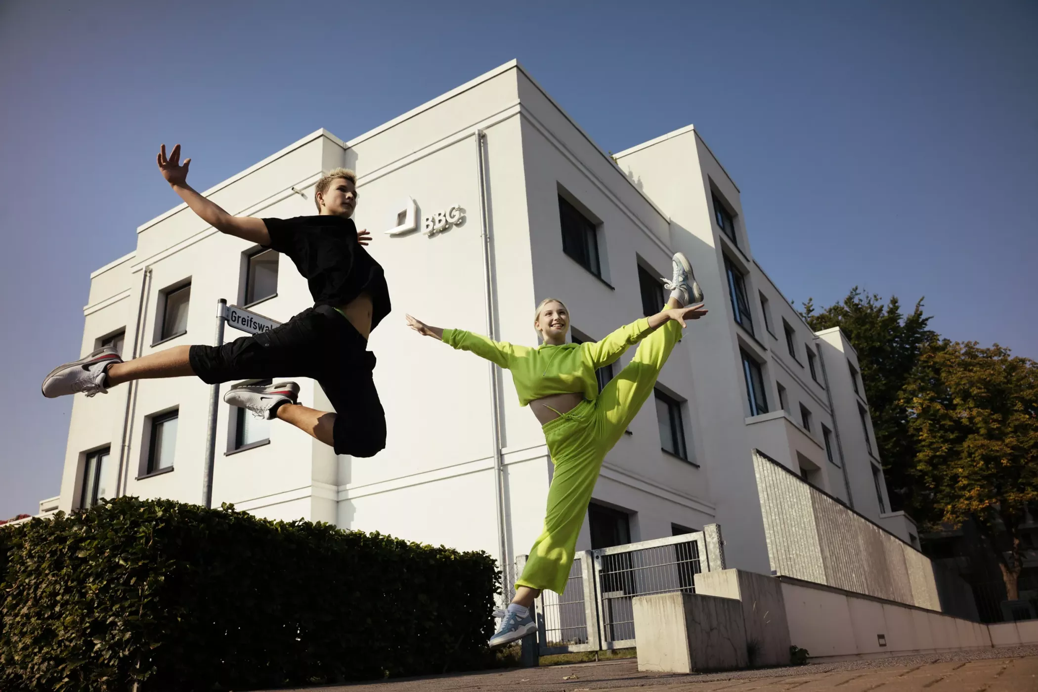 Deux personnes habillées de façon athlétique sautent d'un bâtiment blanc moderne sous un ciel bleu vif.