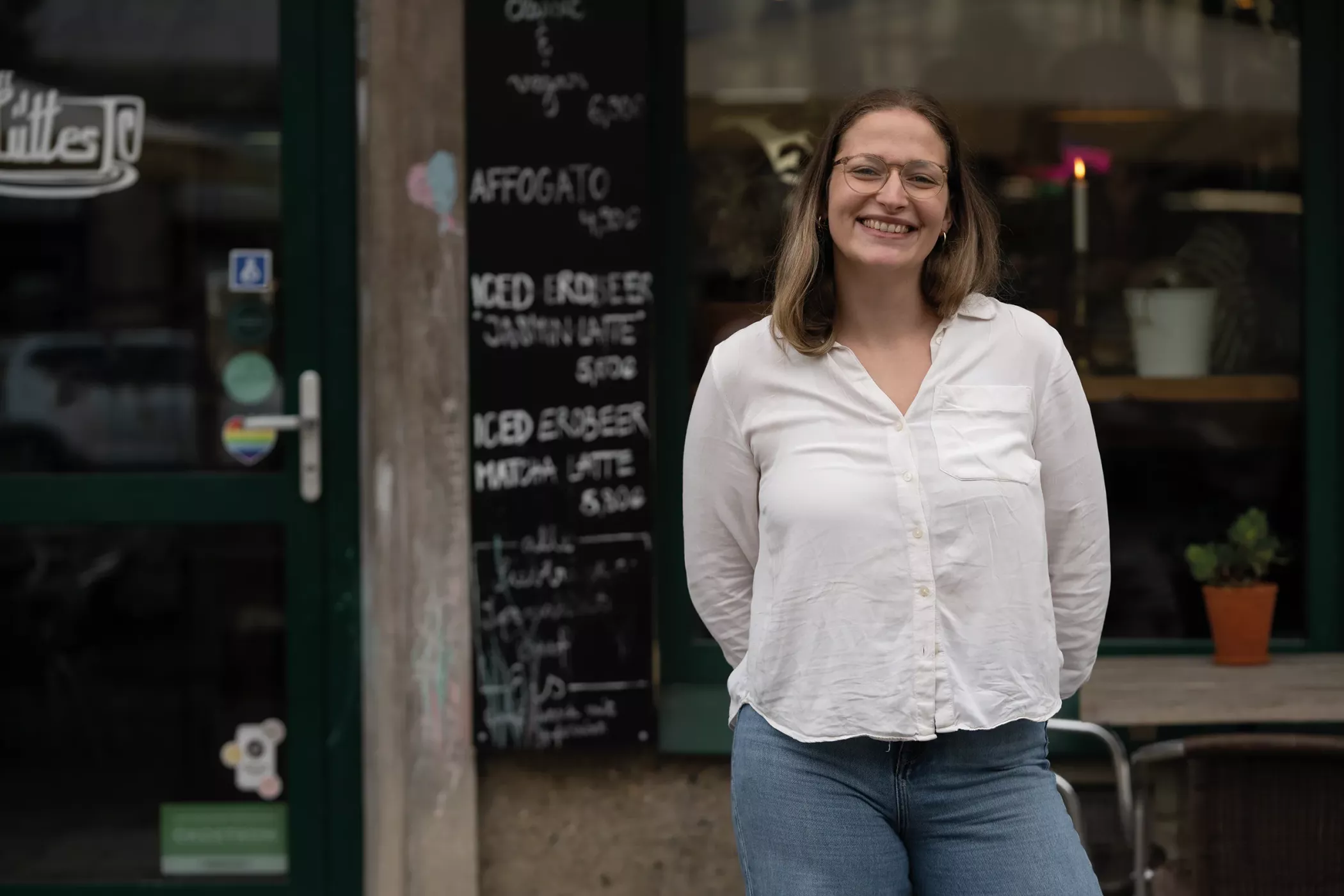 Eine Frau in weißem Hemd und Jeans lächelt, als sie vor einem Café steht, hinter sich eine mit Kreide geschriebene Speisekarte.
