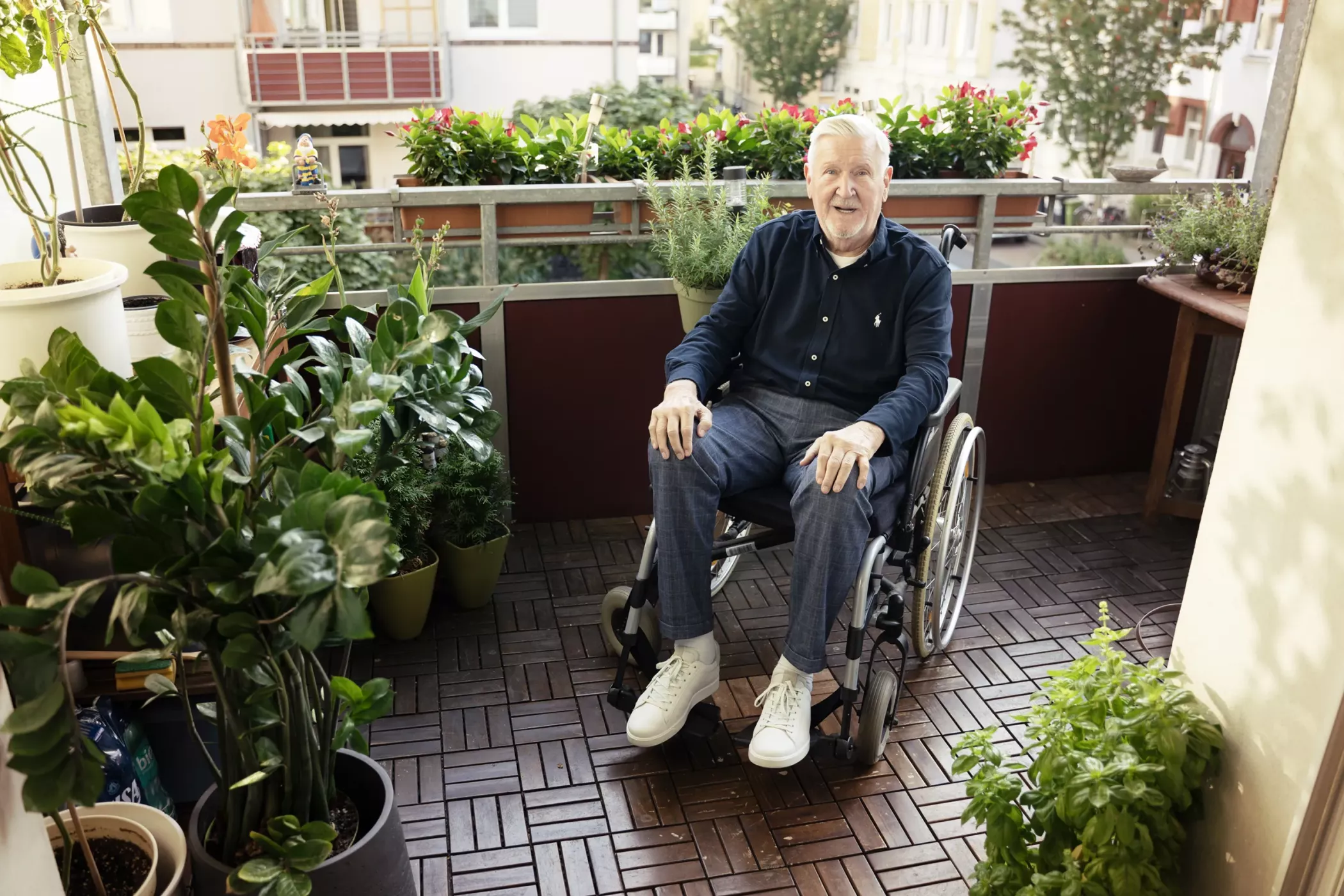 Tekerlekli sandalyedeki yaşlı bir adam saksı bitkileriyle çevrili bir balkonda oturuyor ve kameraya gülümsüyor.