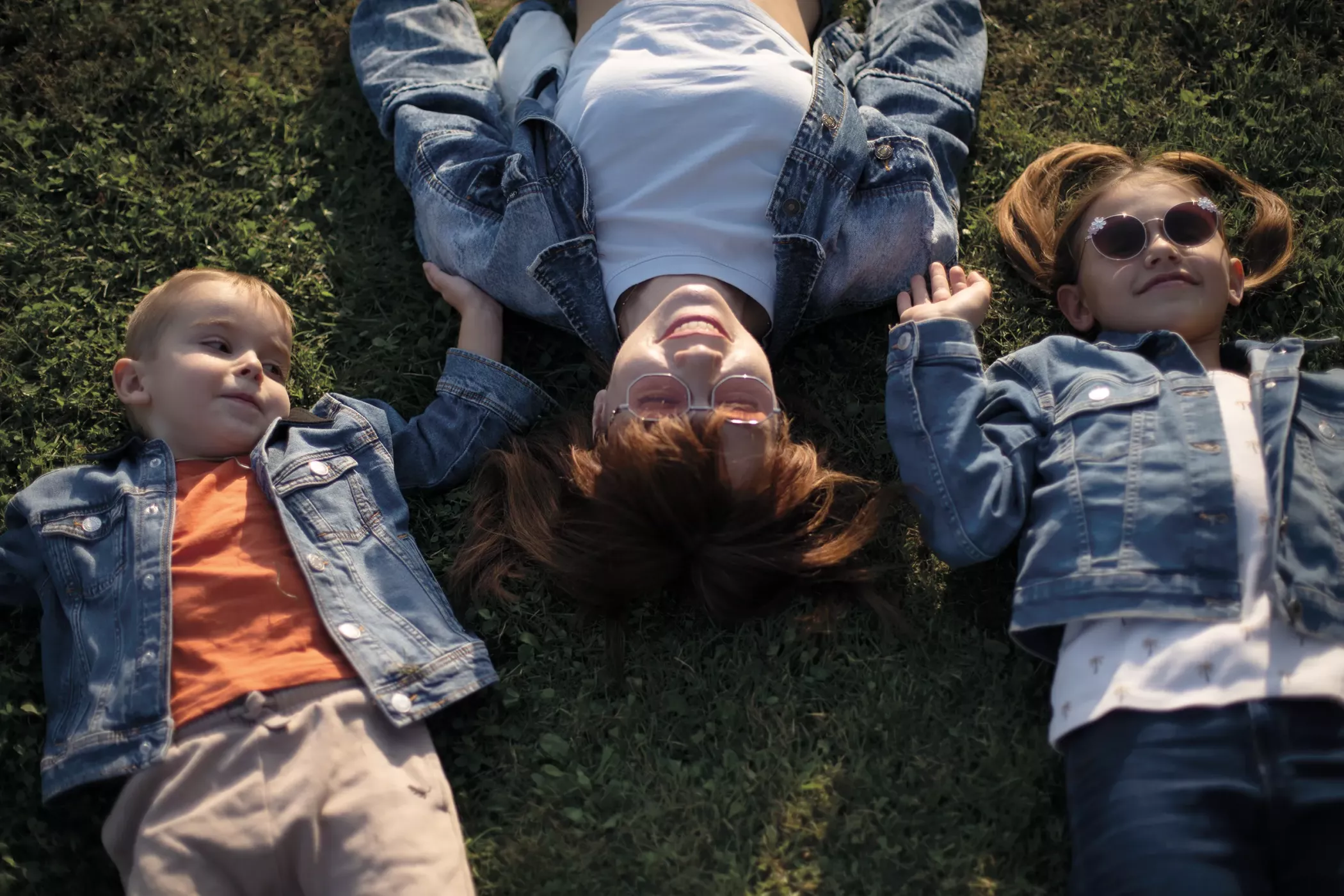 Drei Personen liegen im Gras, halten sich an den Händen und lächeln; zwei Kinder und ein Erwachsener, alle tragen Jeans und Sonnenbrillen.