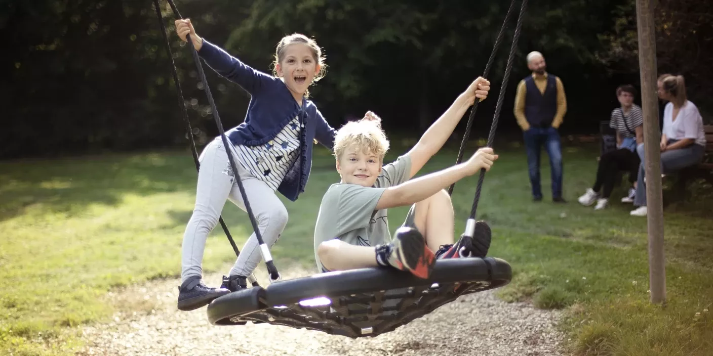 Zwei Kinder schaukeln fröhlich auf einer runden Schaukel in einem Park, während im Hintergrund Erwachsene sitzen und stehen.