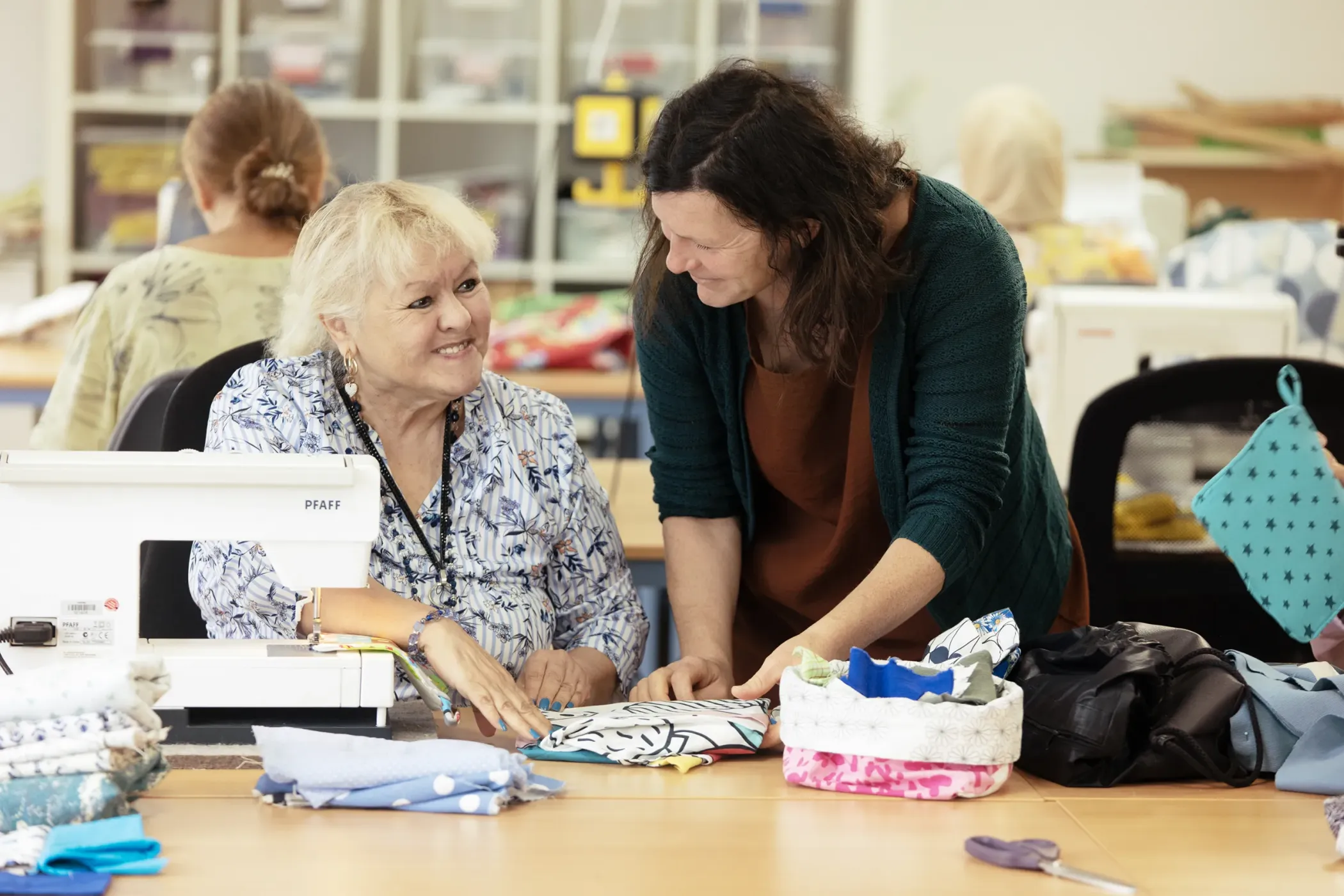 Two women smile and chat as they work on fabrics at a sewing table in a bright co-working space.