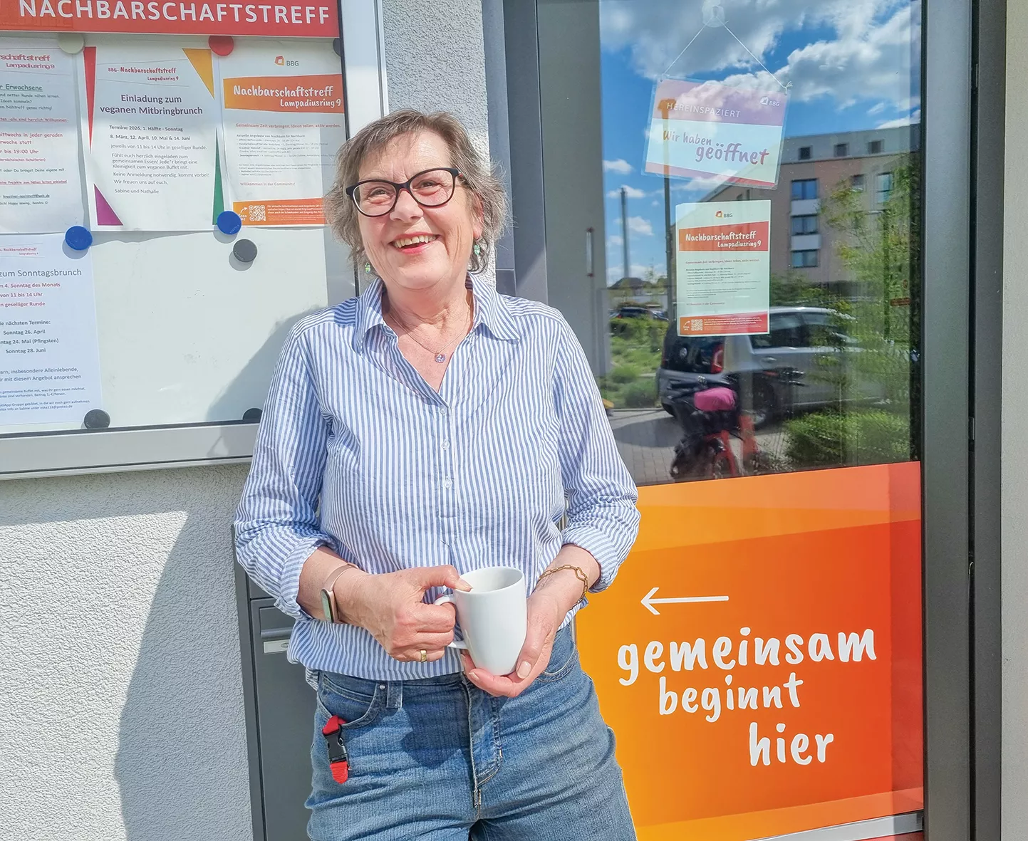 Eine lächelnde Frau mit Brille, die einen Becher in der Hand hält, steht neben einer Tür, an der ein Schild mit der Aufschrift „gemeinsam beginnt hier…“ hängt.