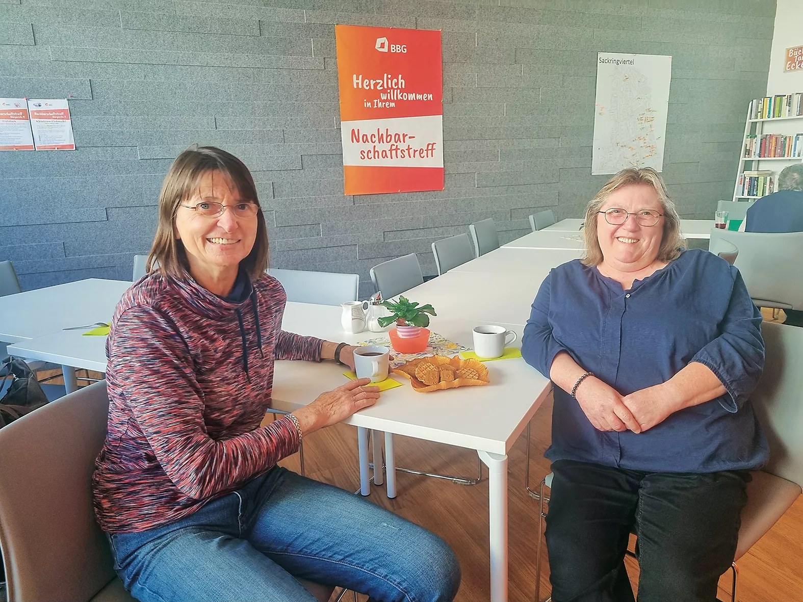 Zwei Frauen sitzen an einem Tisch, auf dem Kaffee, Brot und Obst stehen, und lächeln in einem gemütlichen, hellen Gemeinschaftsraum, dessen Wände mit Plakaten geschmückt sind.