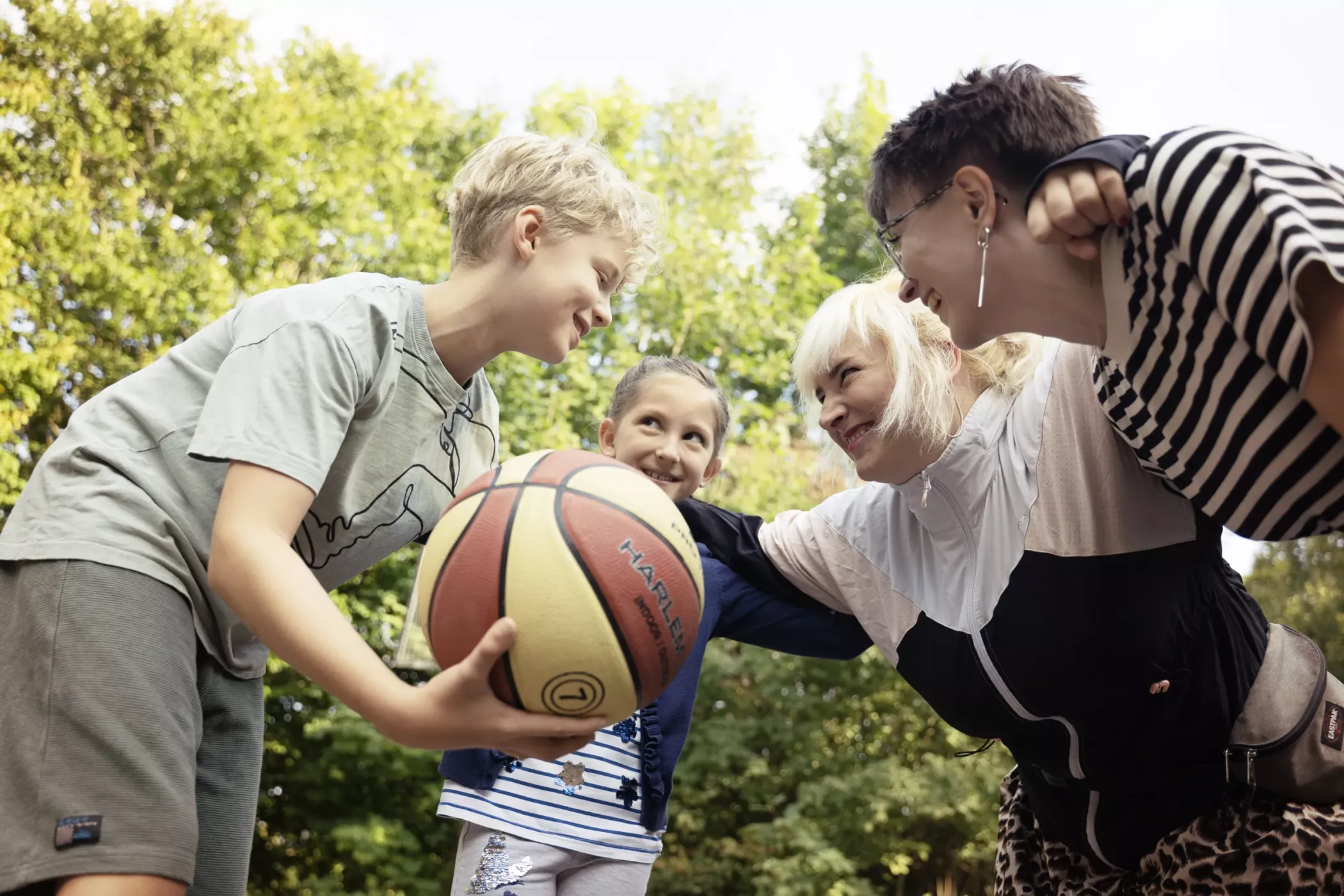 Vier Personen, darunter zwei Kinder und zwei Erwachsene, versammeln sich im Freien und lächeln, während sie einen Basketball halten, mit Bäumen im Hintergrund.