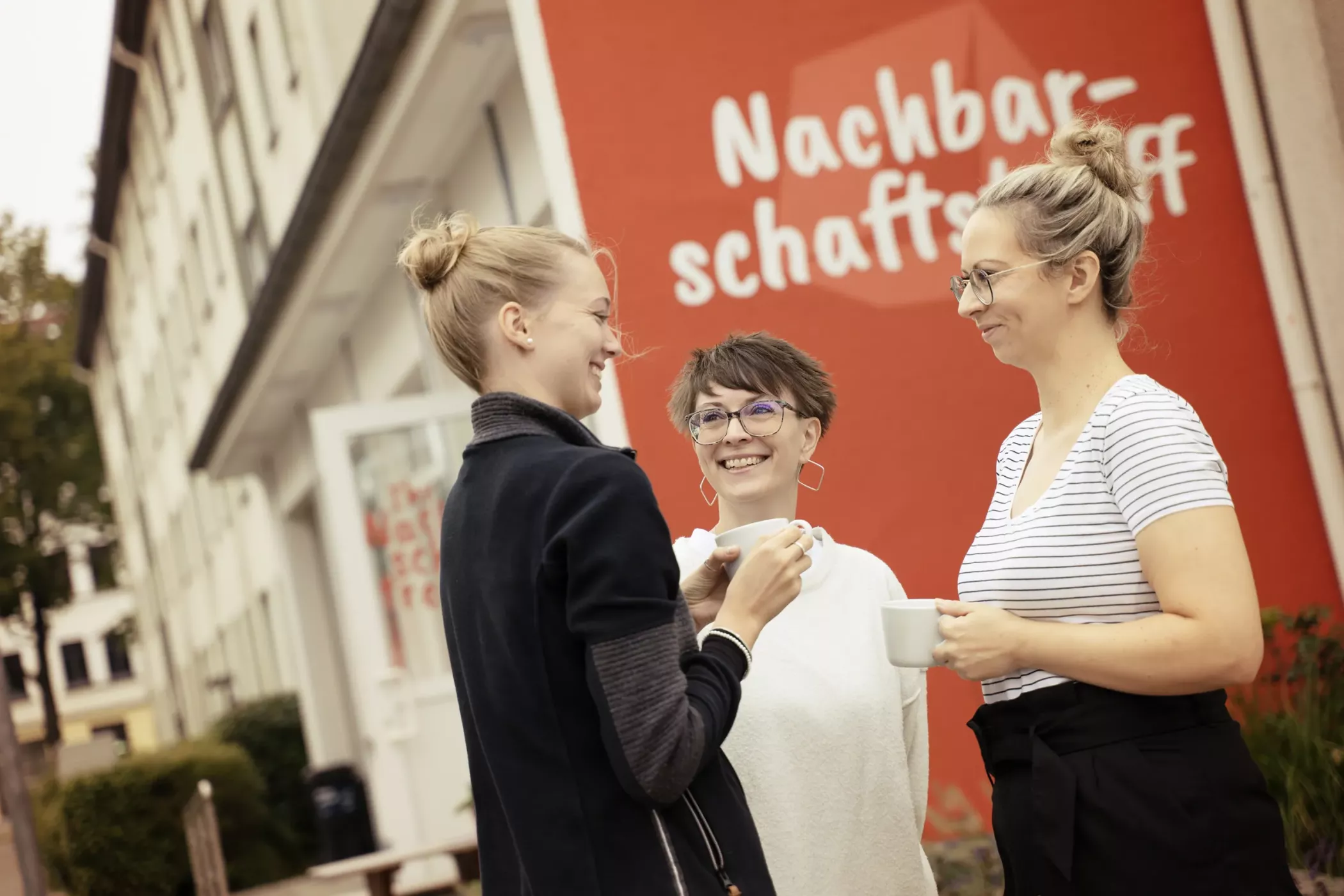 Drei Frauen stehen draußen, lächelnd und plaudernd, mit Kaffeetassen in der Hand, vor einer roten Wand mit deutscher Aufschrift.