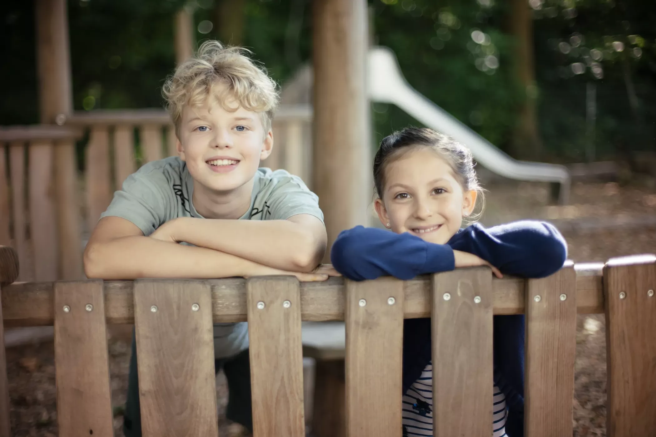 Zwei lächelnde Kinder lehnen an einem Holzzaun auf einem Spielplatz mit Bäumen und einer Rutsche im Hintergrund.
