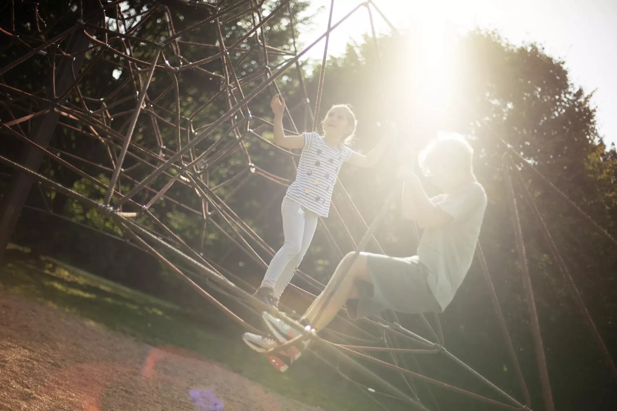 İki çocuk, arkalarında güneş parlarken oyun alanındaki bir ip iskeleye tırmanıyor.