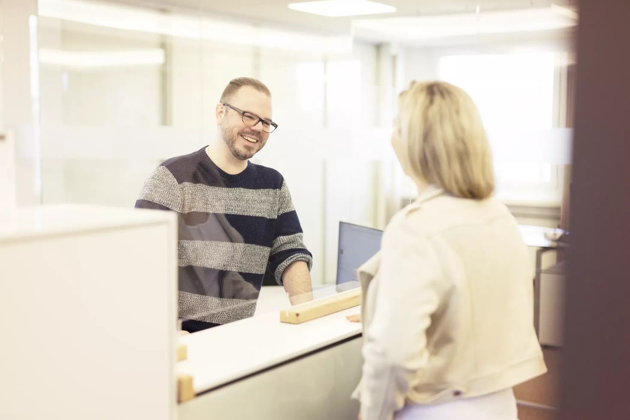 A man with glasses, smiling and talking to a woman at a table in a bright, modern office.