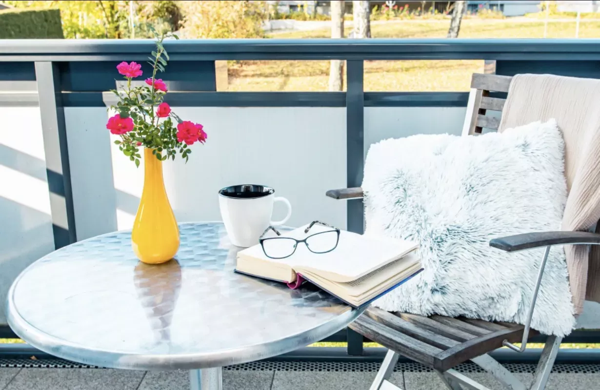 Eine gemütliche Terrassenszene mit einem Stuhl, einem flauschigen Kissen, einem runden Tisch, einer gelben Vase mit rosa Blumen, einem Buch, zwei Gläsern und einem Kaffeebecher.