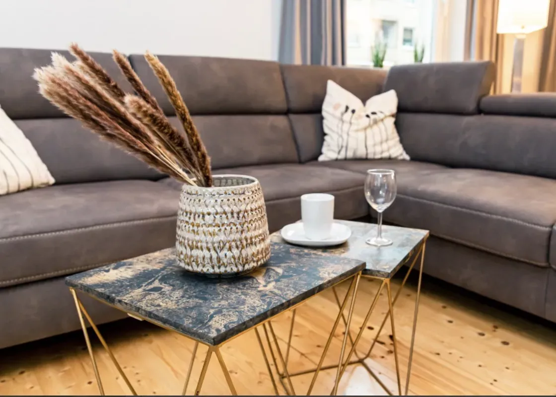Ein modernes Wohnzimmer mit einem grauen Sektionssofa, Kissen und zwei Marmortischen, auf denen eine Vase mit getrockneten Kräutern, ein Glas und ein Weinglas stehen.