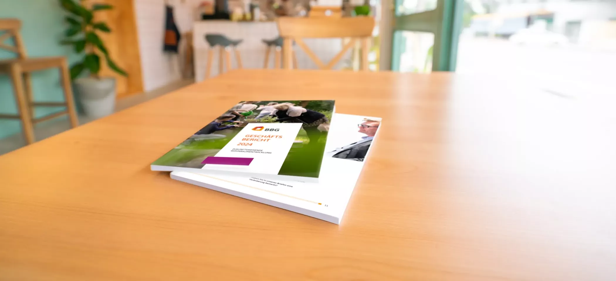 Eine Broschüre auf einem Holztisch in einem hellen Raum mit Stühlen, Pflanzen und einem Fenster im Hintergrund.
