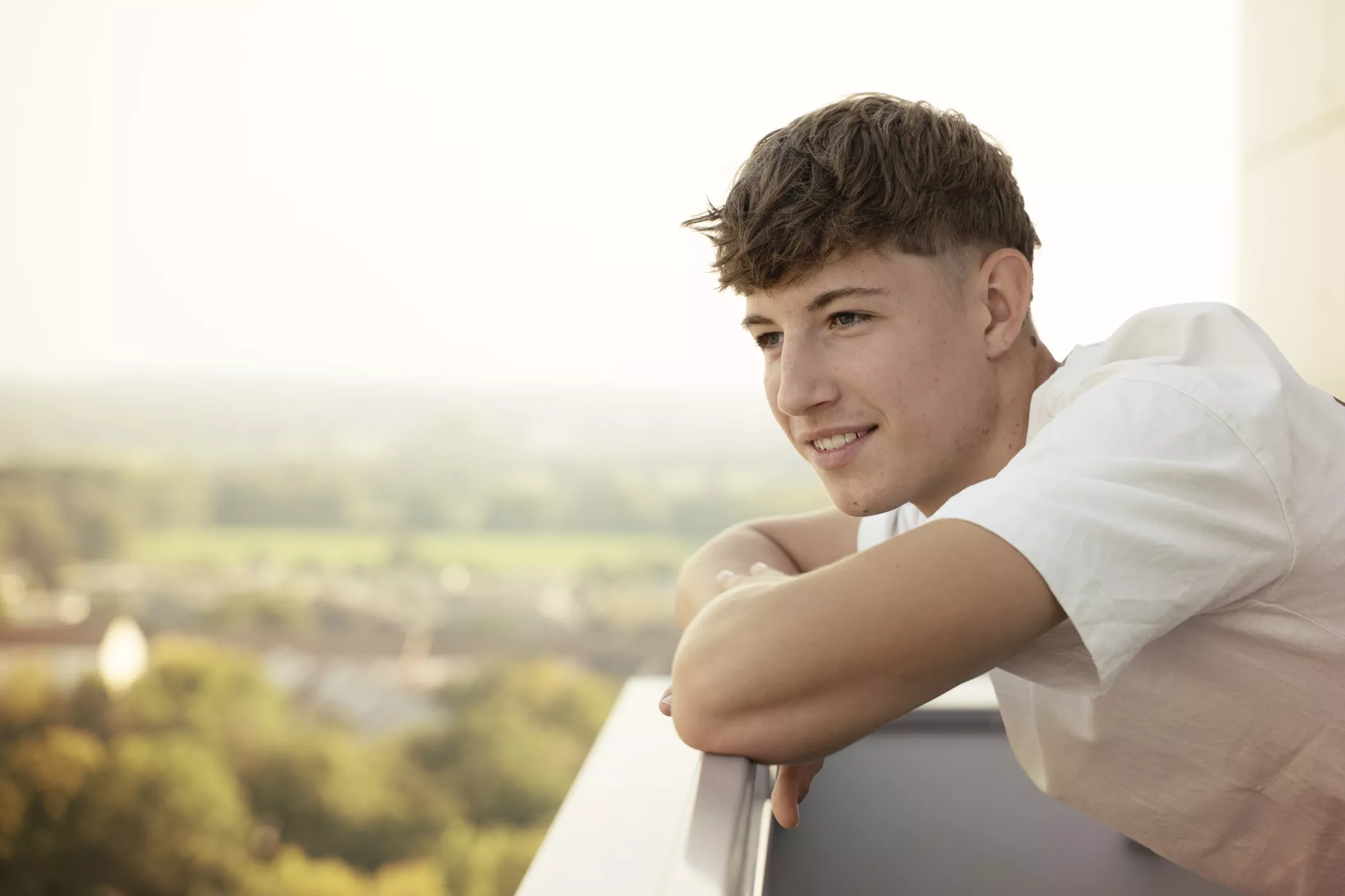 Beyaz gömlekli genç bir adam balkonun korkuluklarına yaslanmış, gülümsüyor ve güneşli bir günde bulanık bir manzarayı düşünüyor.