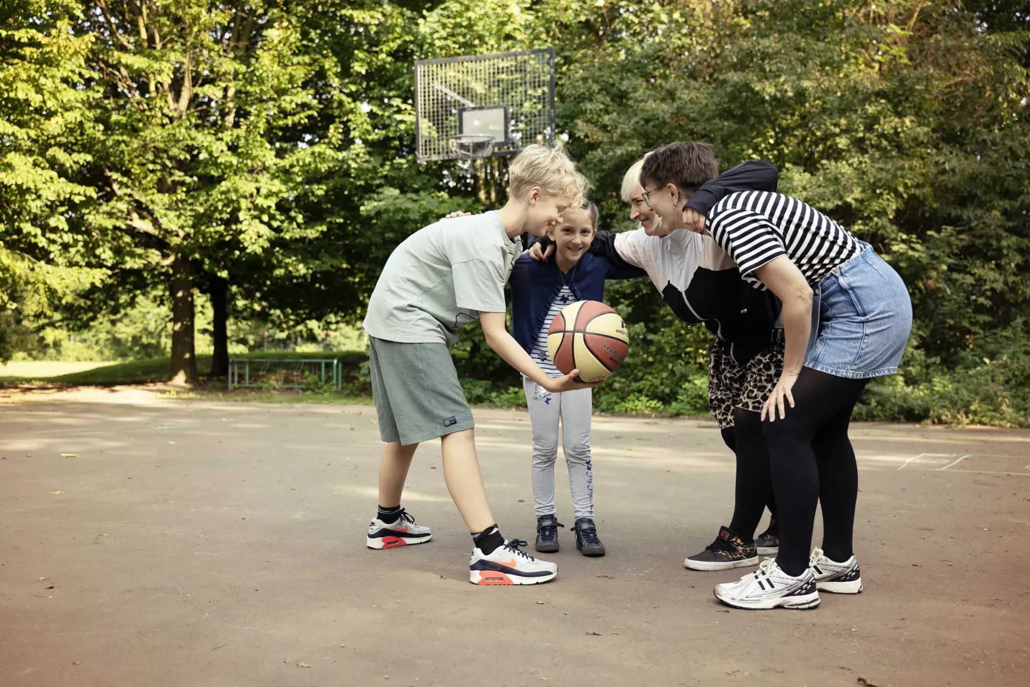 Zwei Kinder und zwei Erwachsene im Freien, lächelnd und um einen Basketball auf einem Spielplatz herum, mit Bäumen und einem Basketballkorb im Hintergrund.