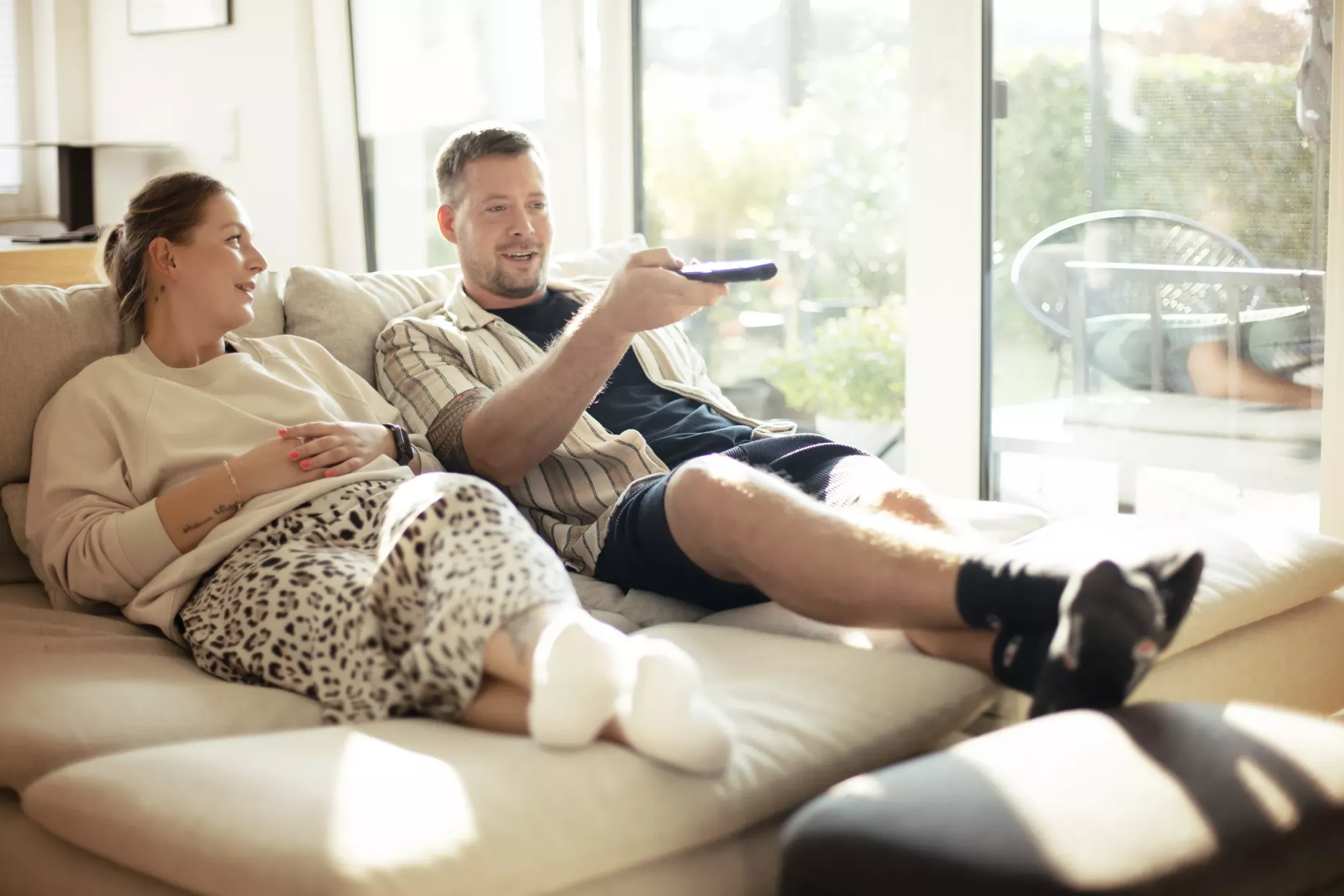 Bir çift kanepede dinlenmekte, sohbet etmekte ve büyük pencereli aydınlık oturma odasında televizyon izlemektedir.