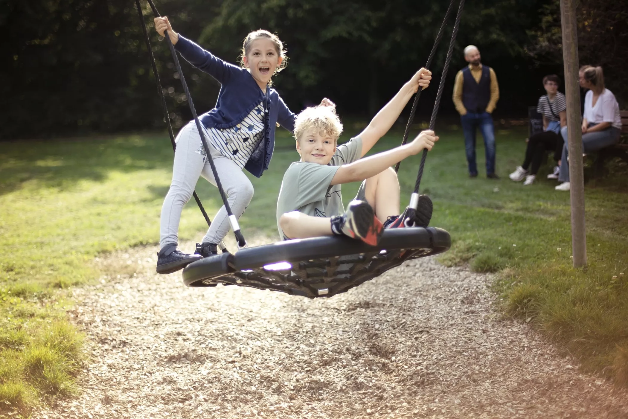 Parkta yuvarlak bir salıncakta gülümseyen iki çocuk, arka planda oturan ve ayakta duran yetişkinler.