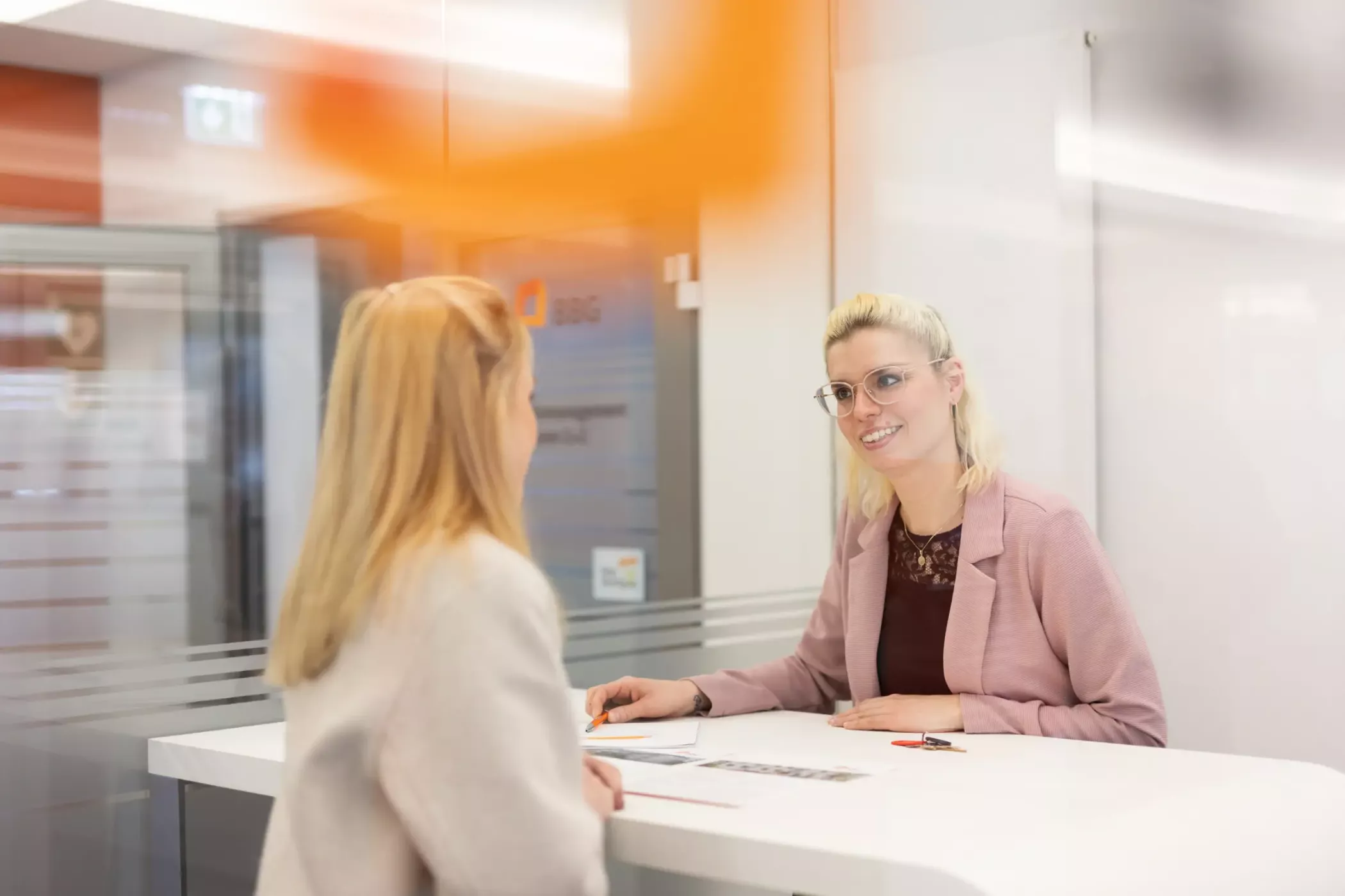 Two women are talking at the reception desk of a modern office; one is sitting and smiling, the other is standing opposite her.
