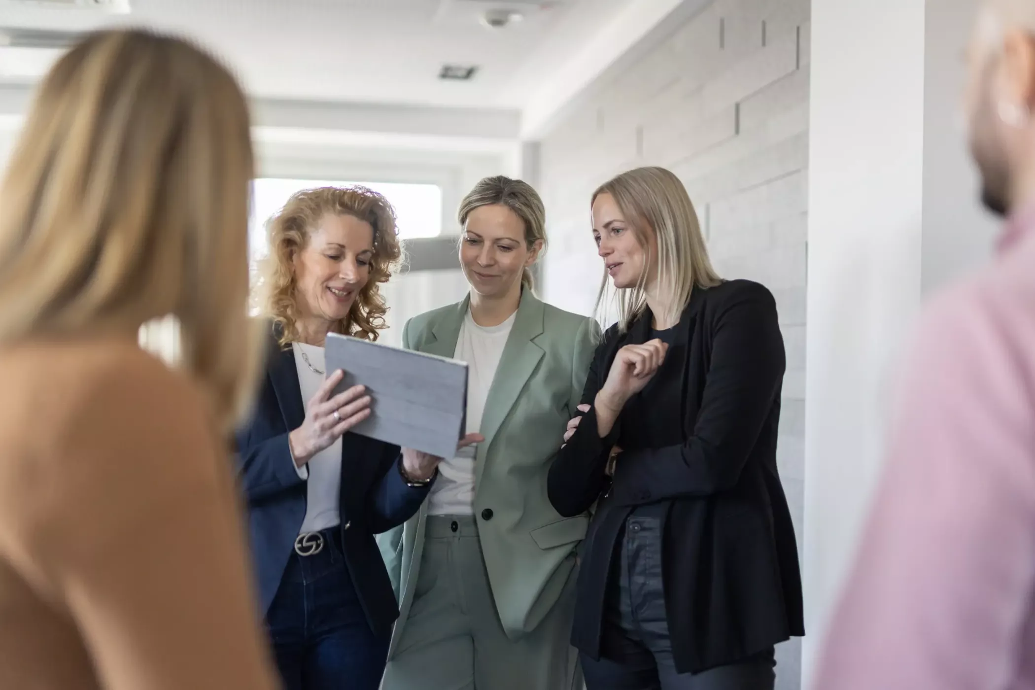 ثلاث نساء يقفن معًا ويبتسمن وينظرن إلى جهاز لوحي، مع وجود شخصين آخرين غير واضحين في المقدمة.