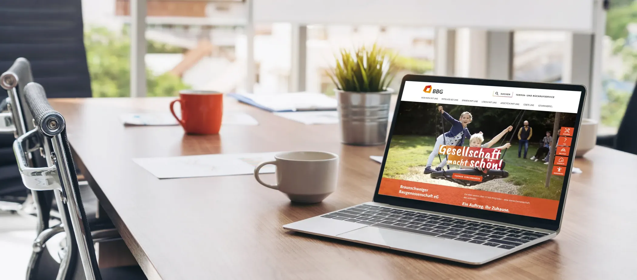 Ein Laptop auf einem Schreibtisch zeigt eine Website an; daneben stehen Kaffeetassen, Papiere, eine Pflanze und ein heller Bürohintergrund.