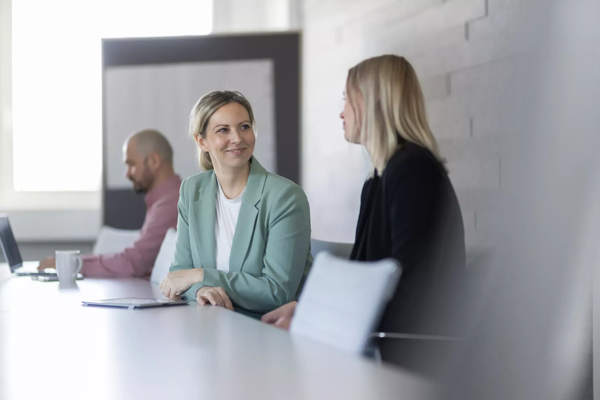 Drei Personen sitzen an einem Konferenztisch; zwei Frauen unterhalten sich im Vordergrund, während ein Mann im Hintergrund an einem Laptop arbeitet.
