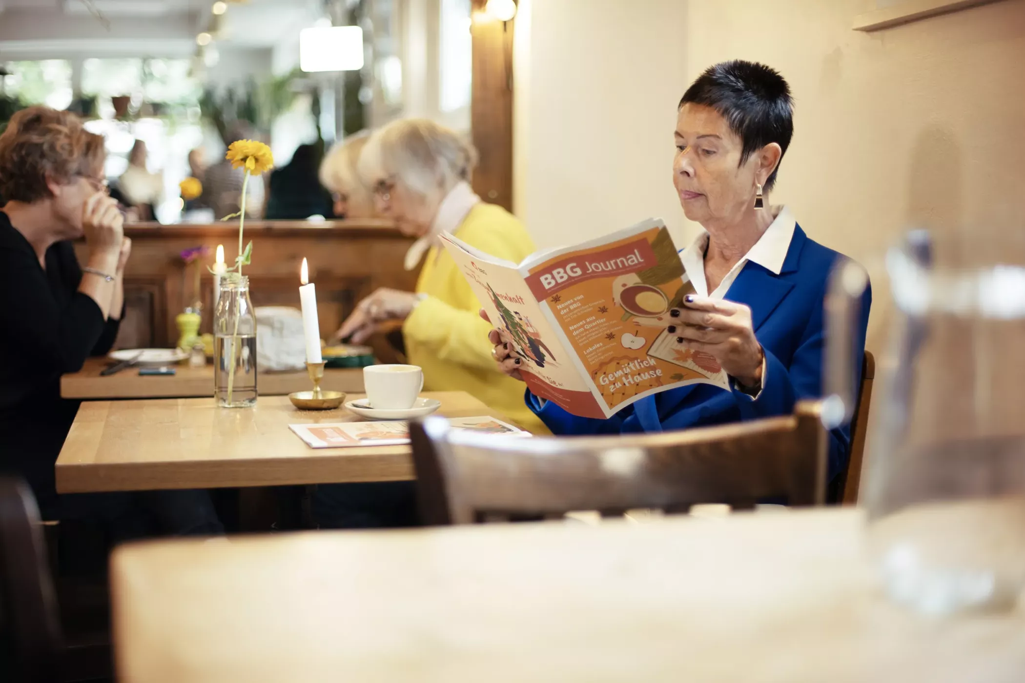 Eine ältere Frau in einer blauen Jacke liest eine Zeitschrift an einem Cafétisch, im Hintergrund sind Kaffee und andere Gäste zu sehen.